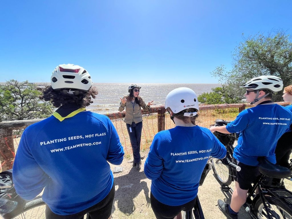 Young people are wearing white helmets and blue t-shirts that say 'Planting seeds, not flags,' the motto of the WeTwo expedition. They're facing outwards looking at the sea and sitting on bikes.