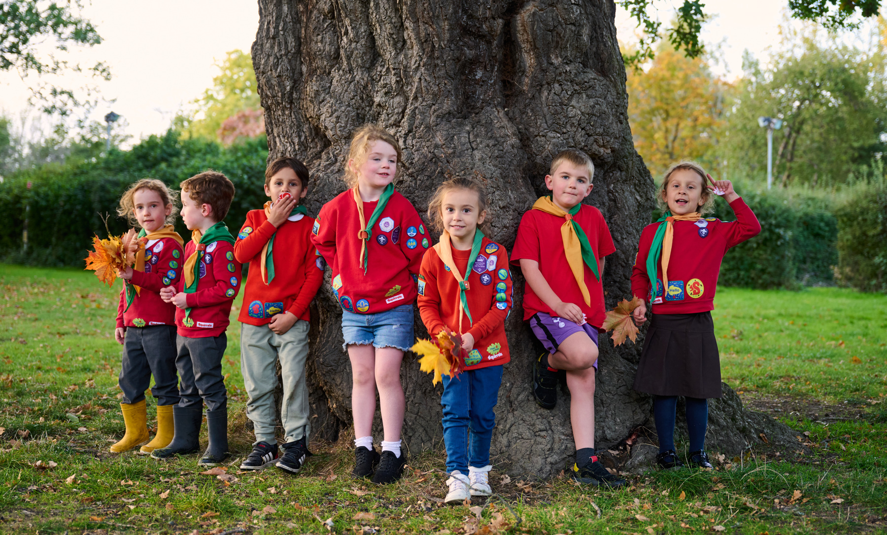 Seven Squirrels in uniform and neckers stand in front of a tree trunk holding leaves in their hands