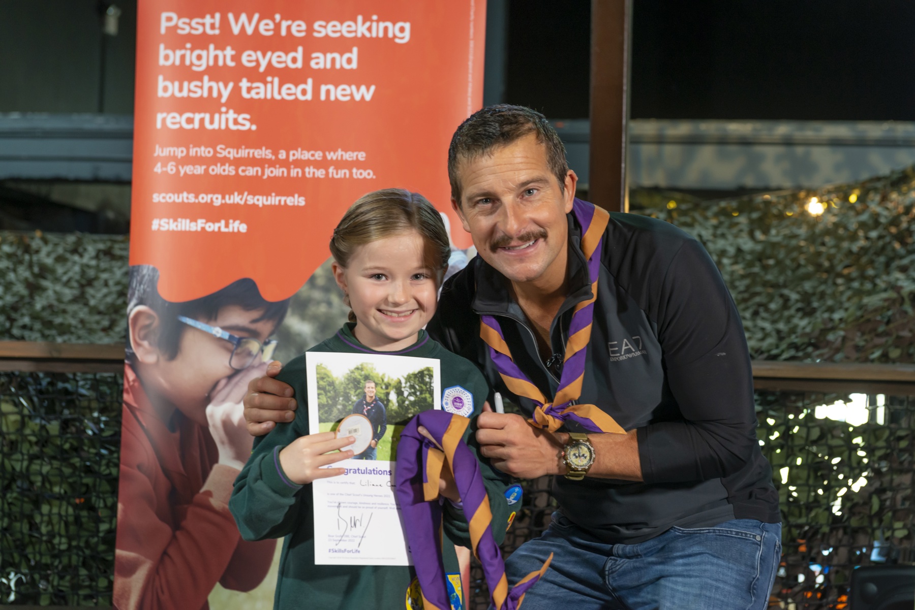 Lilianne is in her cubs uniform and stood with Bear Grylls. They're both smiling. Lilianne is holding her certificate, and Never Give Up badge and necker.