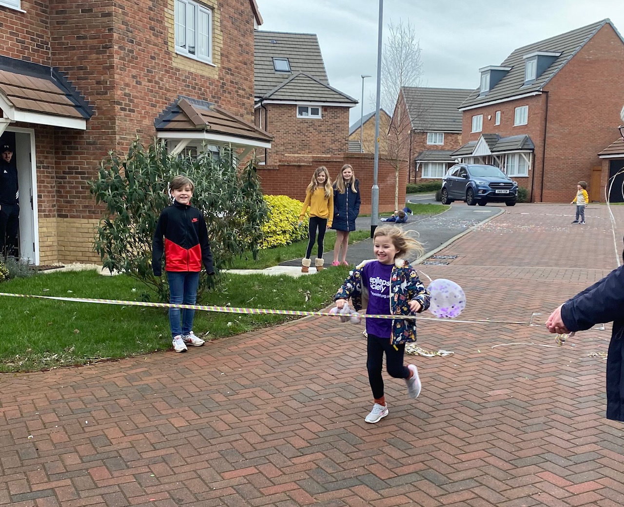 Liliane runs across the finish line, while being watched by friends. She is outside a row of homes and is smiling. She is wearing her purple epilepsy society t-shirt.