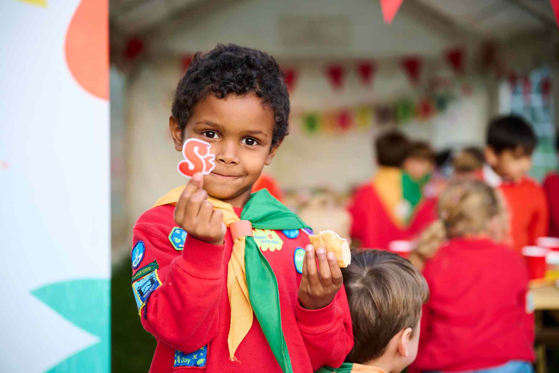 A Squirrel wearing a red jumper smiles at the camera while holding a Squirrels sticker in one hand and a roll in the other.