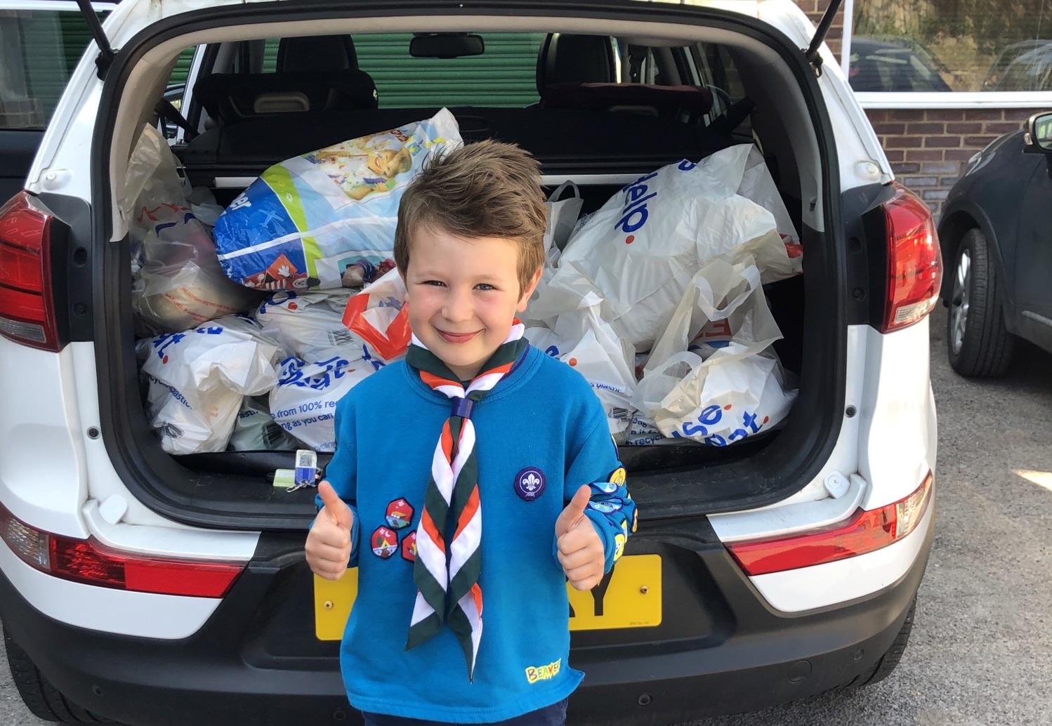 Henry's wearing his Beavers uniform with a necker, holding both his thumbs up and smiling at the camera. He's standing in front of a car boot which has lots of bags of shopping in.