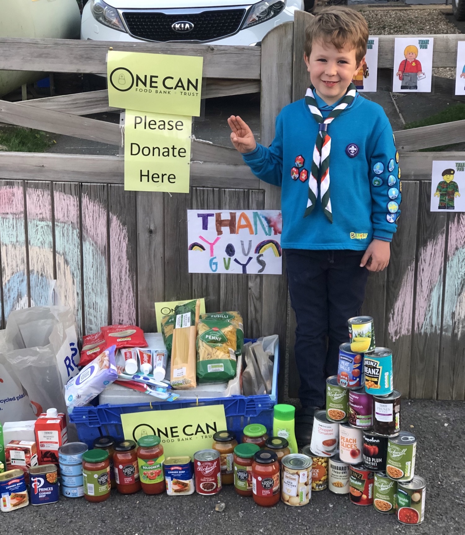 A Beaver in uniform with badges and a necker is holding his hand in the Scout salute in front of a fence. A sign behind him says 'Please Donate Here' and in front of him are lots of cans and tins of long-life food.