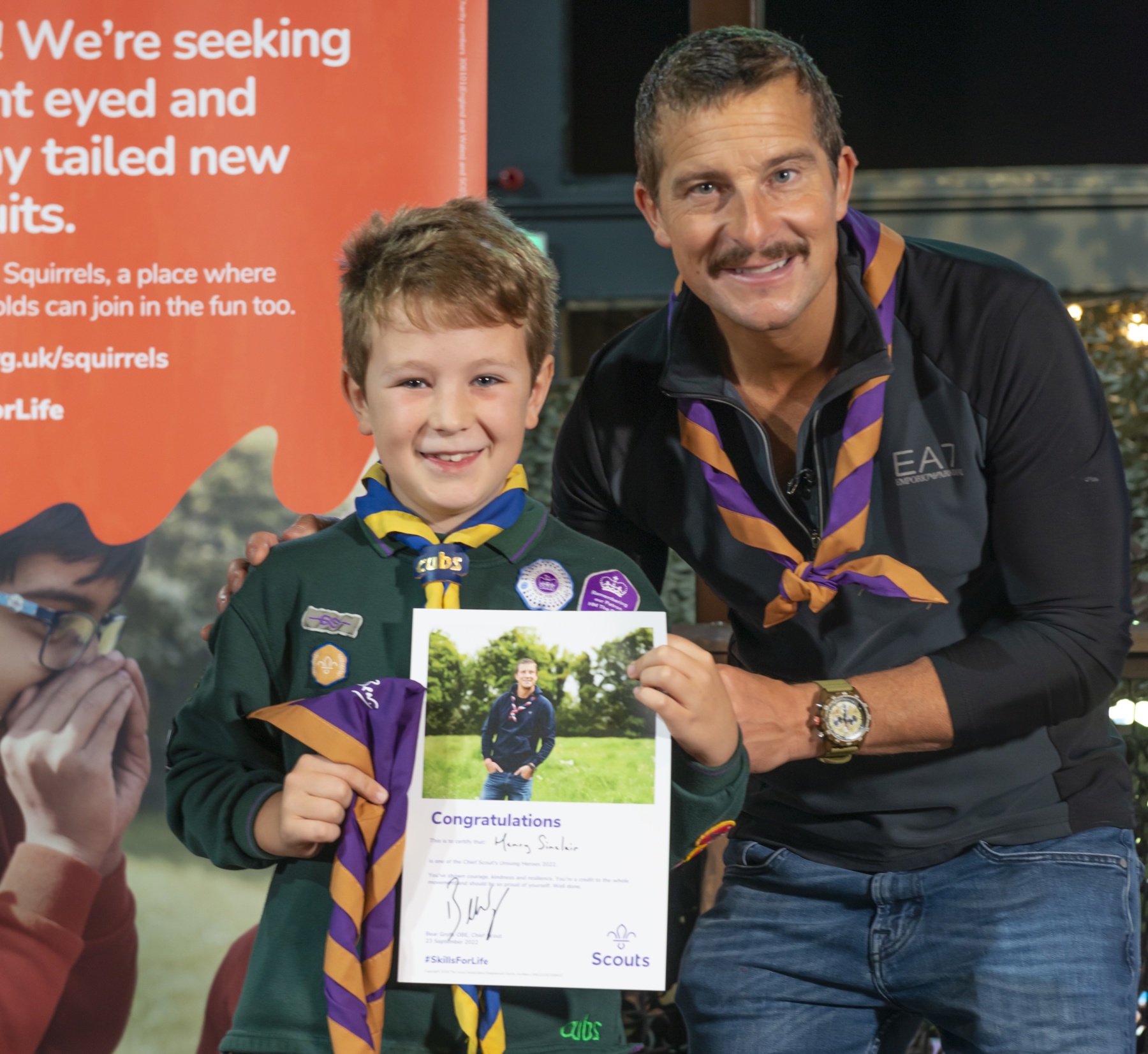 Henry's wearing his Cub uniform and a necker, and is holding a 'Congratulations' certificate. He stands next to Chief Scout Bear Grylls, with both of them smiling at the camera.