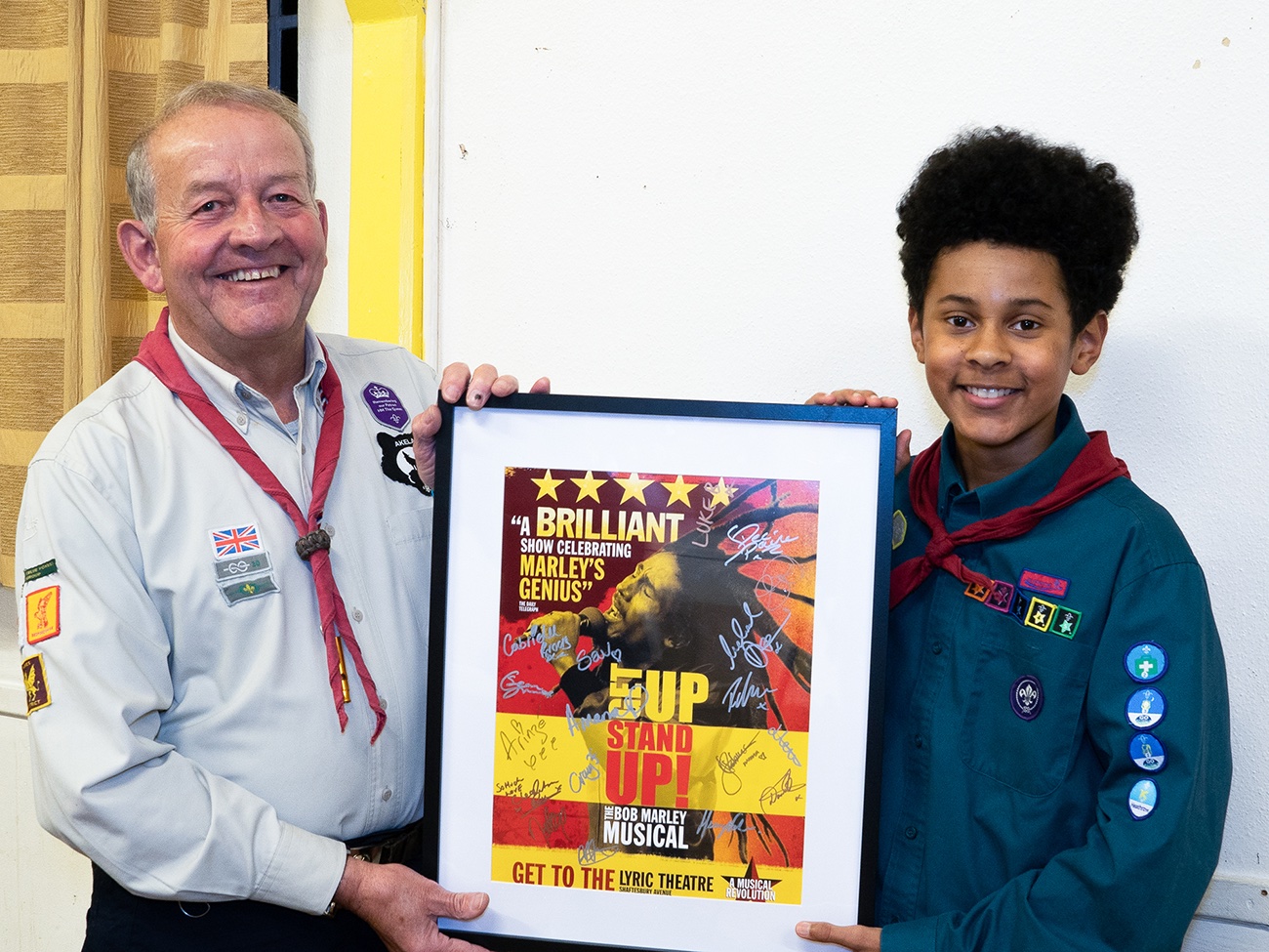 A Cub leader wearing uniform and a red necker is holding a poster of the musical 'Get Up, Stand Up' in a black frame. To the right of the poster is Cub, Maxwell, in uniform and a red necker. They're both smiling at the camera.