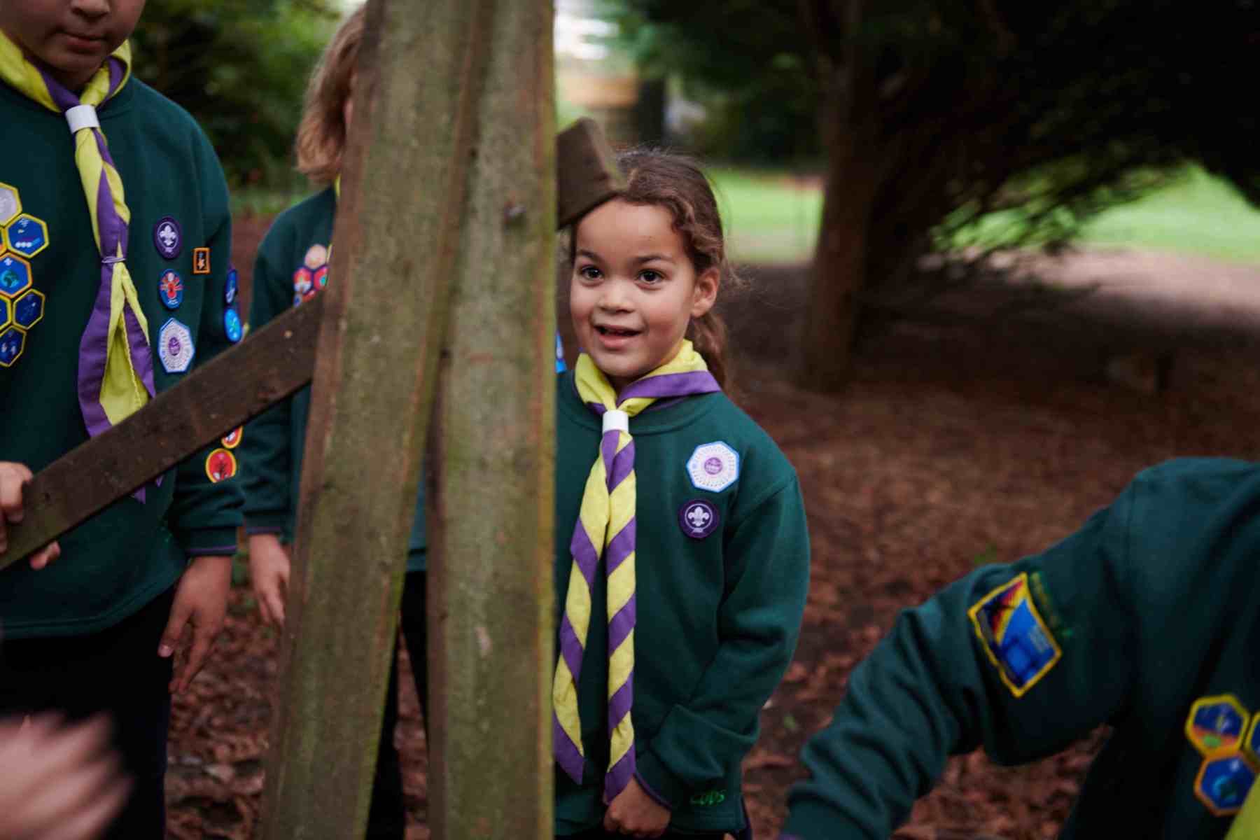 A Cub wearing a green uniform  and yellow and purple necker looks straight ahead at some wood planks.