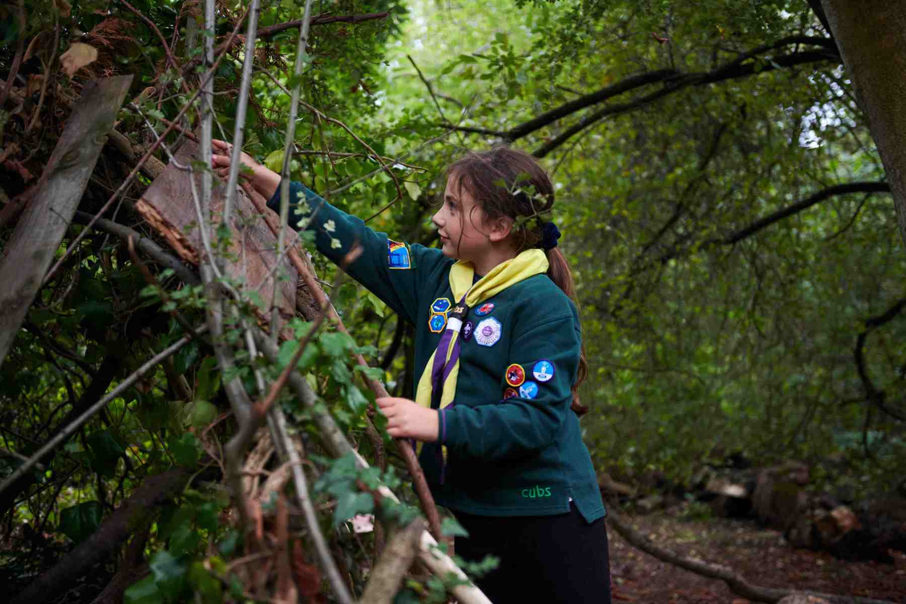 A girl is building a Den in a wooded area, reaching out and holding sticks. She's a cub with a yellow necker on.