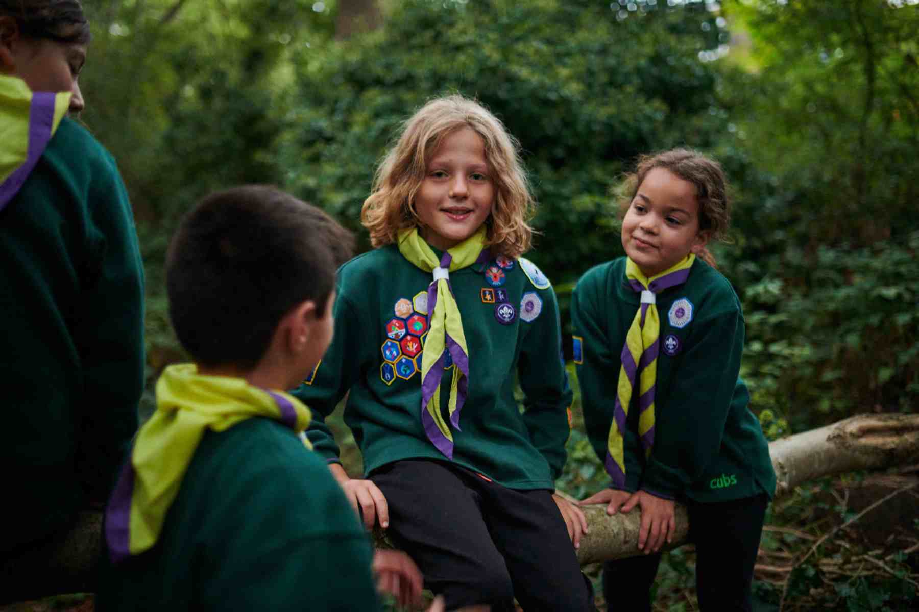 Two smiling Cubs in green uniforms and neckers talking to each other while sitting on the branch of a tree.