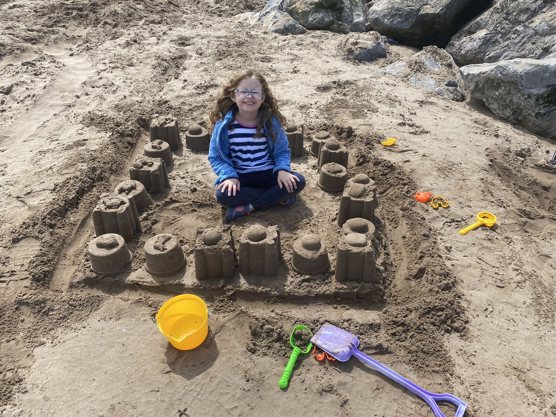 Ella is at the beach and sat in the middle of a big sandcastle, smiling.