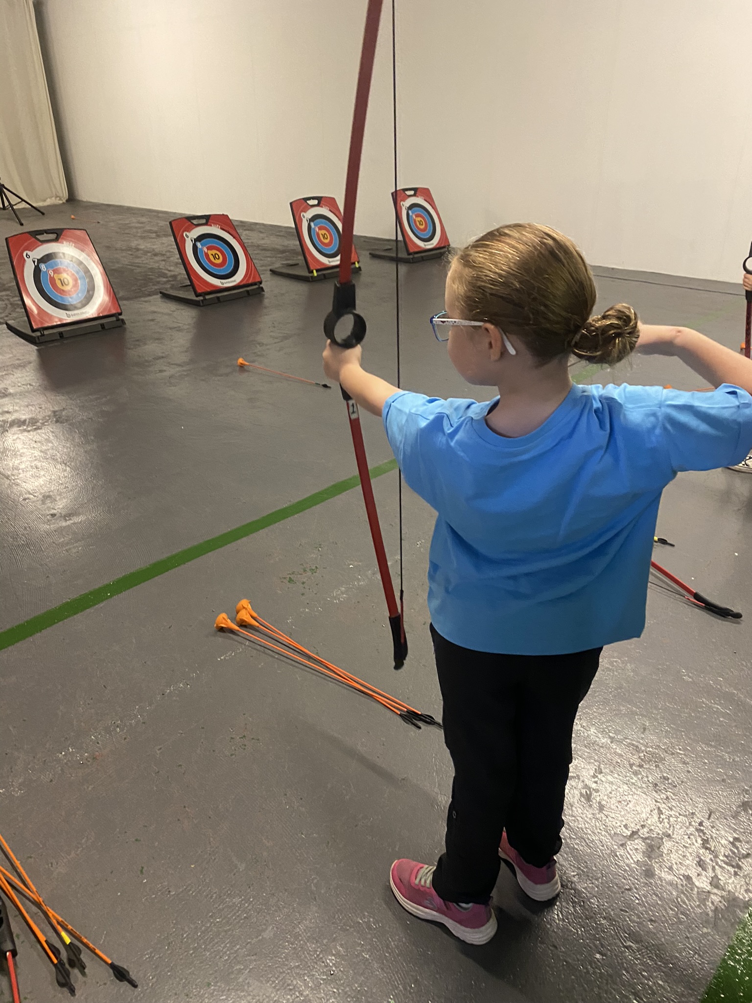 Ella is trying archery. She is stood indoors while holding a bow and soft archery arrow. The bow's pointed at the target and Ella is about to let go of the arrow.