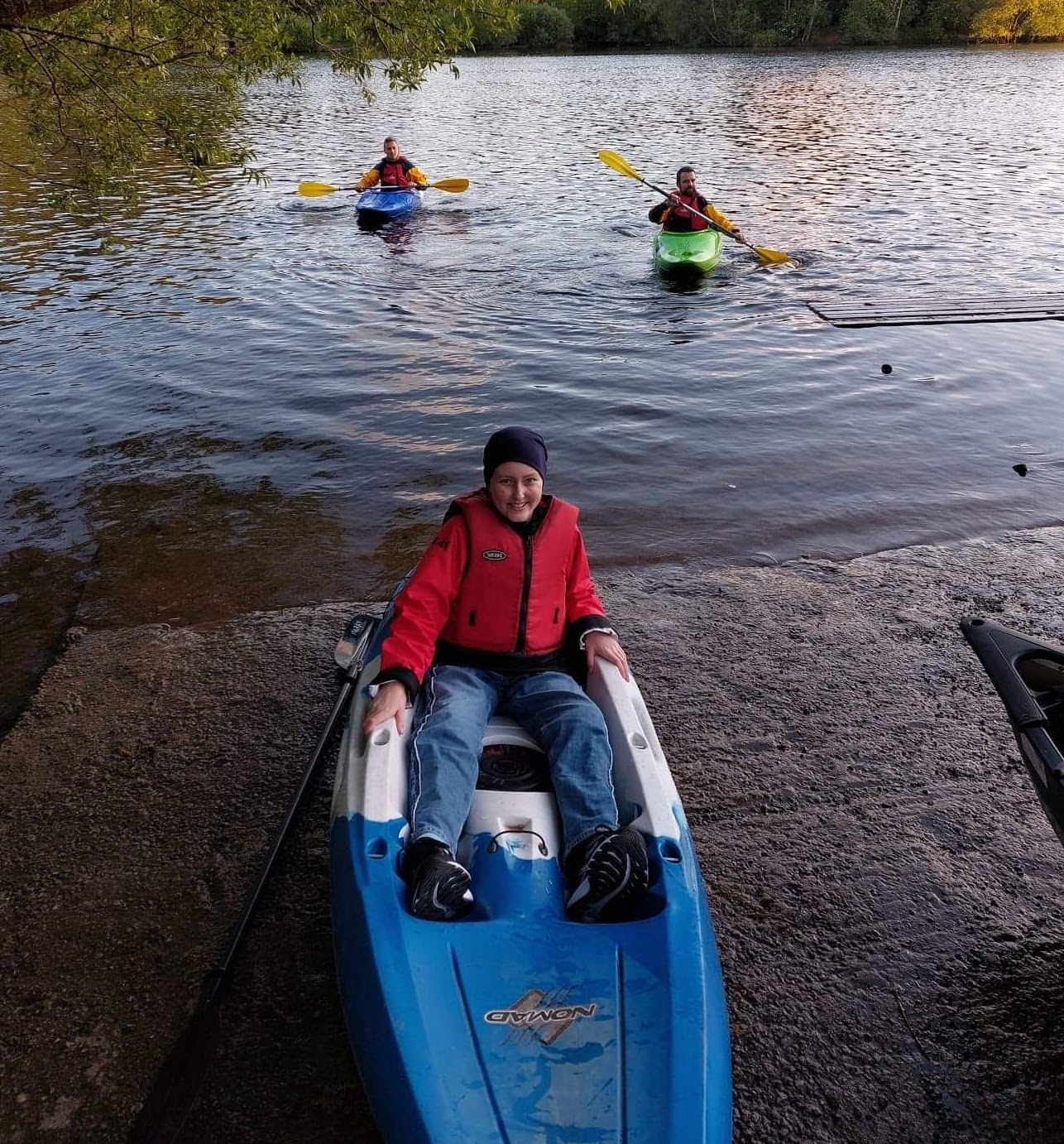 The image shows Isabelle sat in a kayak on concrete near a lake. There are two people in kayaks on the water behind her, with trees in the background.