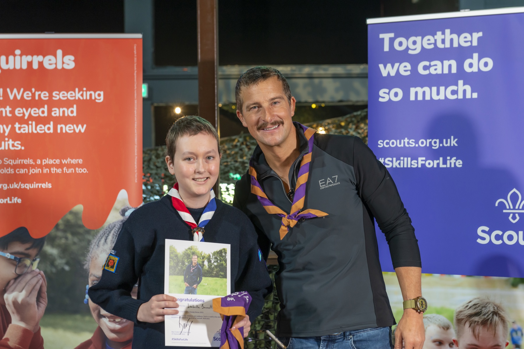 Isabelle is wearing her uniform and necker, holding a 'congratulations' certificate and an orange and purple 'never give up' necker. She's stood next to Bear Grylls, who's wearing an orange and purple necker, and they're both smiling at the camera.