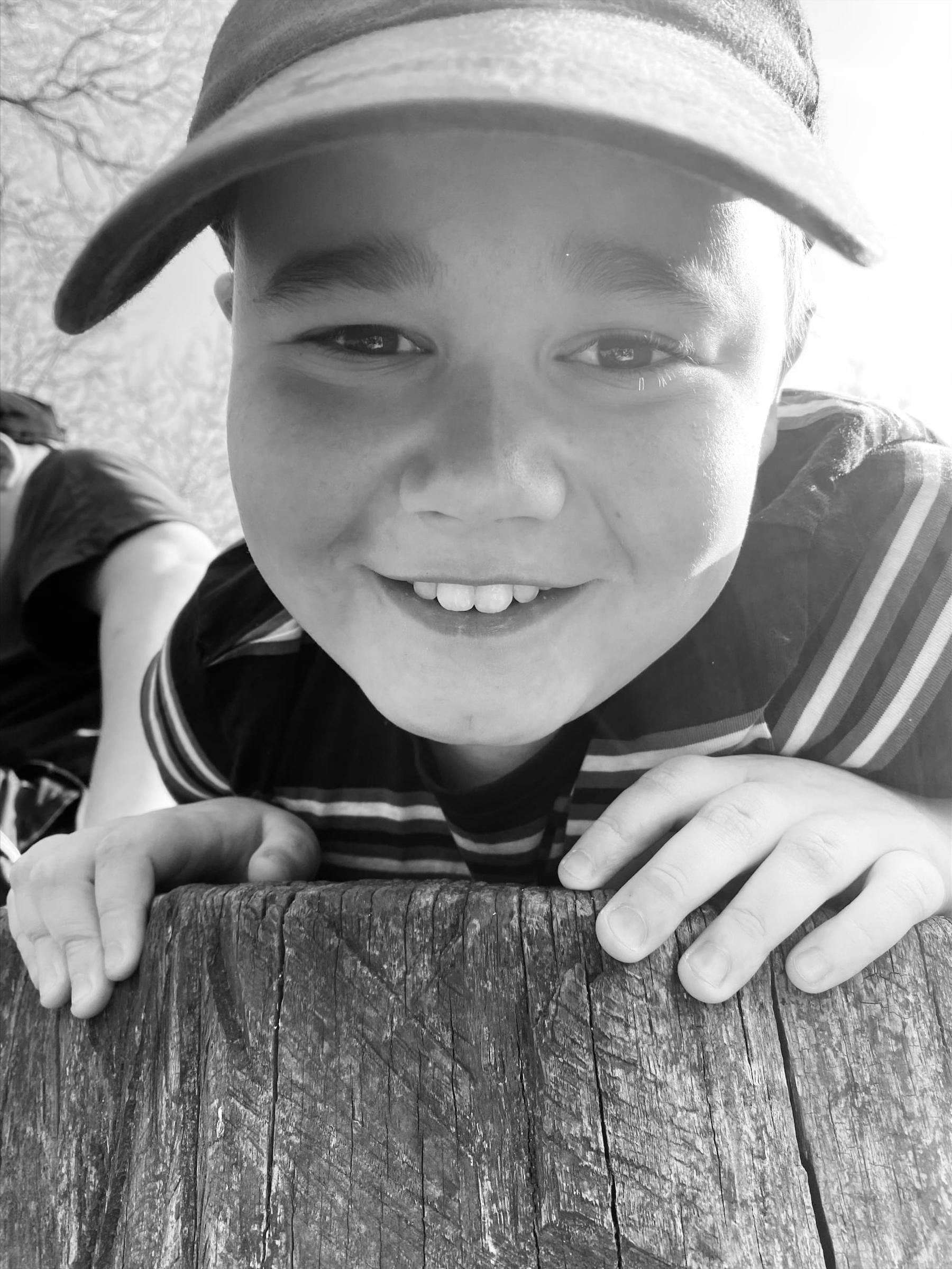 Ben is smiling at the camera. He is wearing a hat and striped t shirt. He's leaning on a fence post.