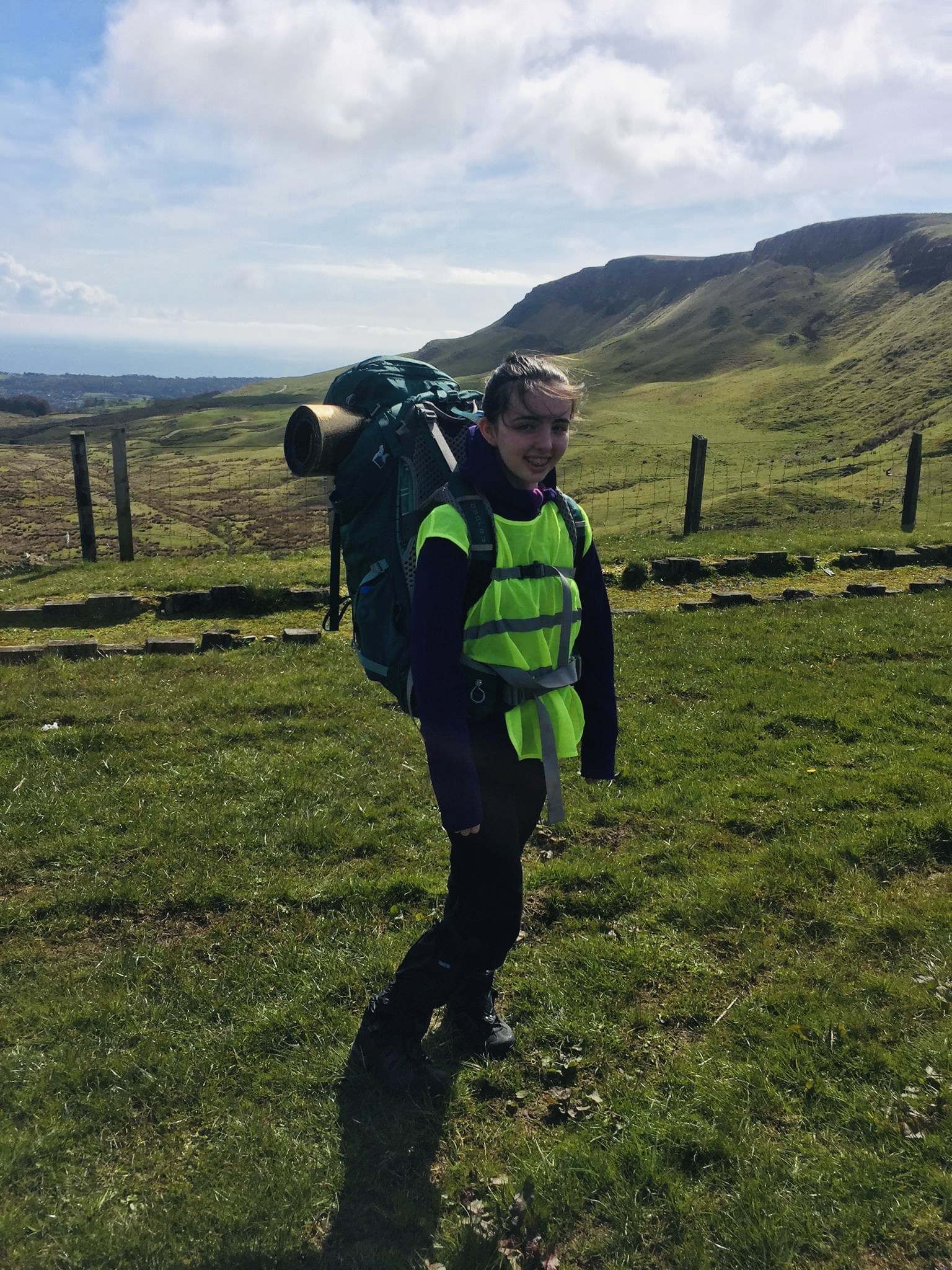 Nicole has a backpack on her back and she's standing in a field with grassy hills behind her.