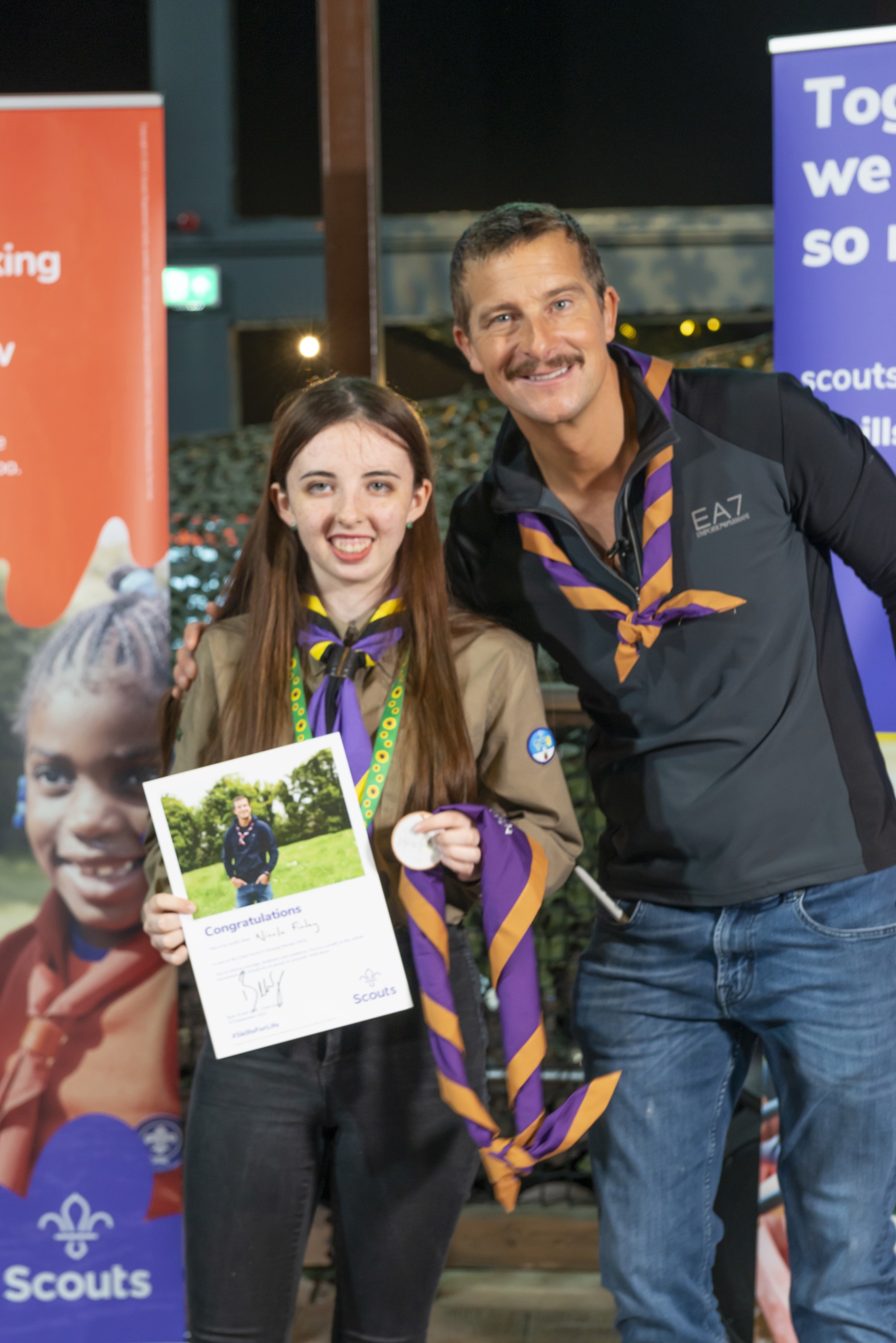 Nicole is holding her Unsung Heroes certificate standing next to Chief Scout Bear Grylls. They're both wearing neckers and smiling at the camera.