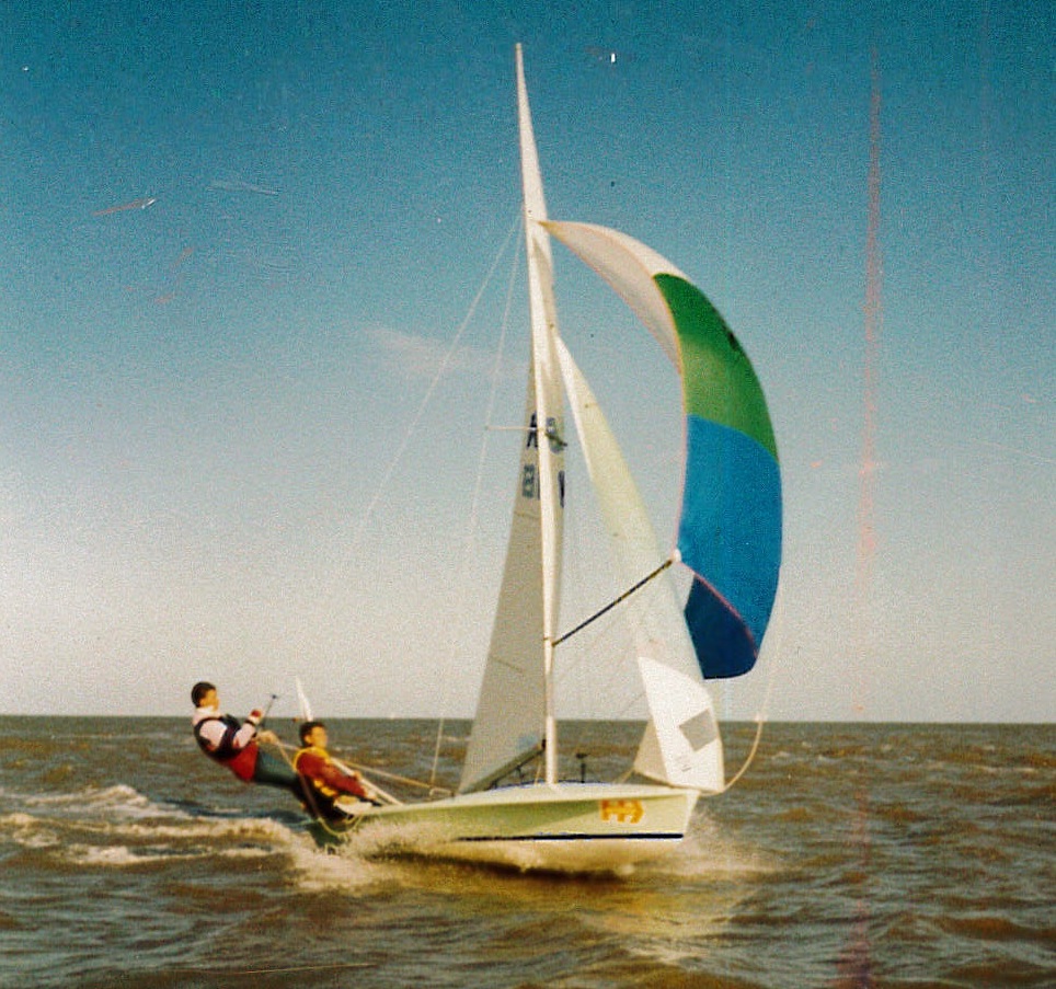 Mike is holding onto his boat's sail on the side of his rowing boat in the middle of the sea, with another sailor sat beneath him. The wind is pushing the green, white and blue sail out to the right.