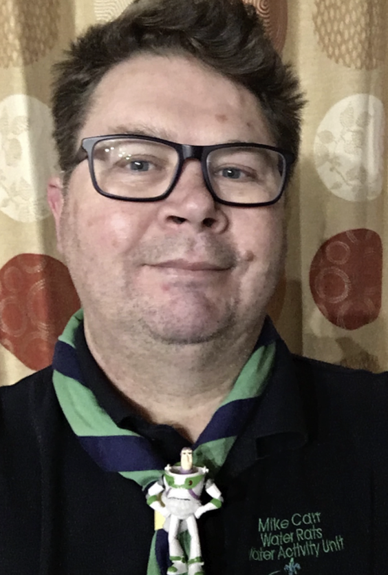 Mike is looking at the camera and wearing black glasses. He's in his Scouts Water Activity Unit uniform and wearing a necker with a Buzz Lightyear figure on.