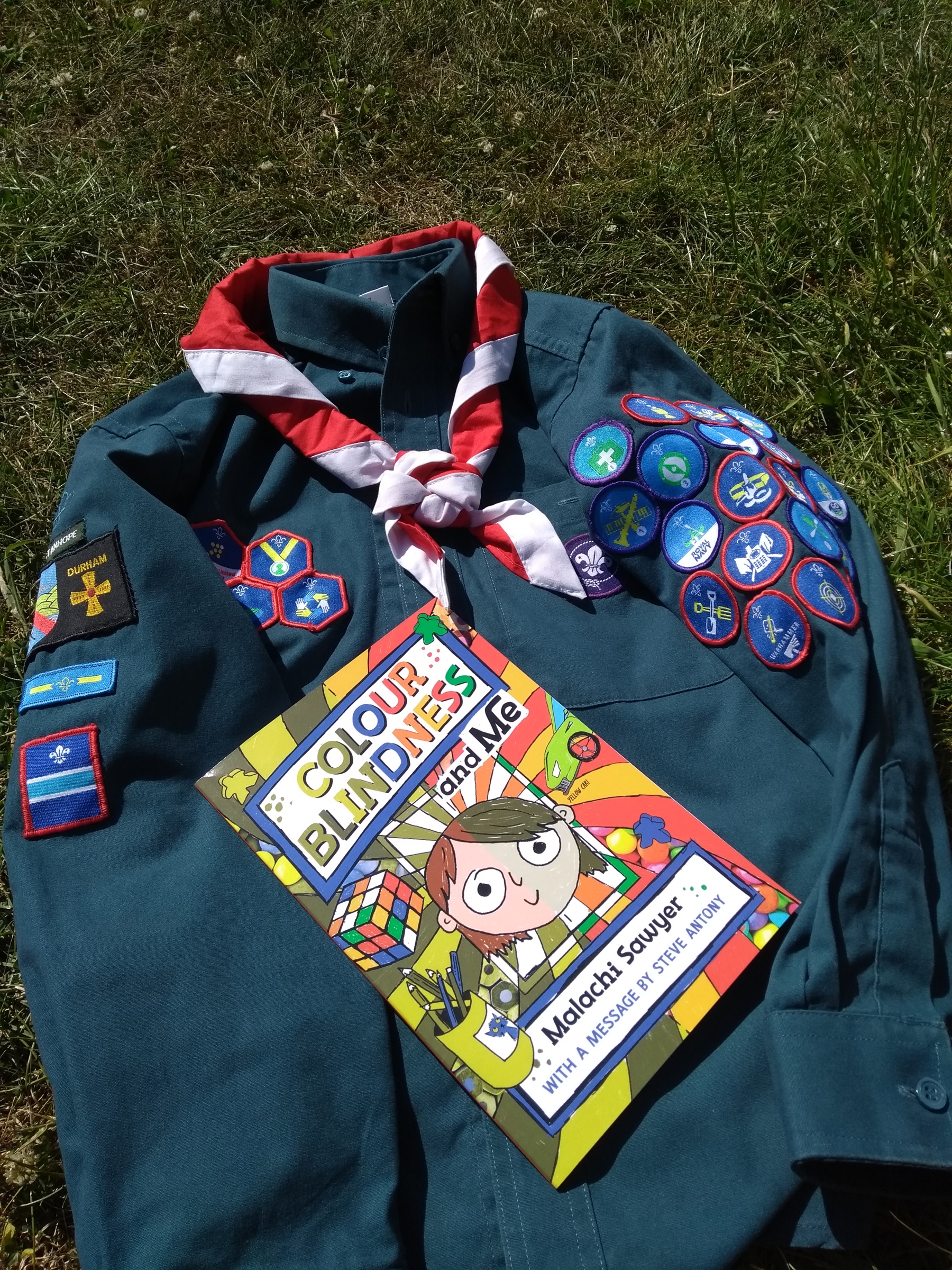 A Scout shirt is laid out on grass with a red and white necker around the colour. There are lots of badges sewn onto the shirt and the book by 12-year-old Malachi, 'Colour Blindness and Me,' is laying on top of the shirt.