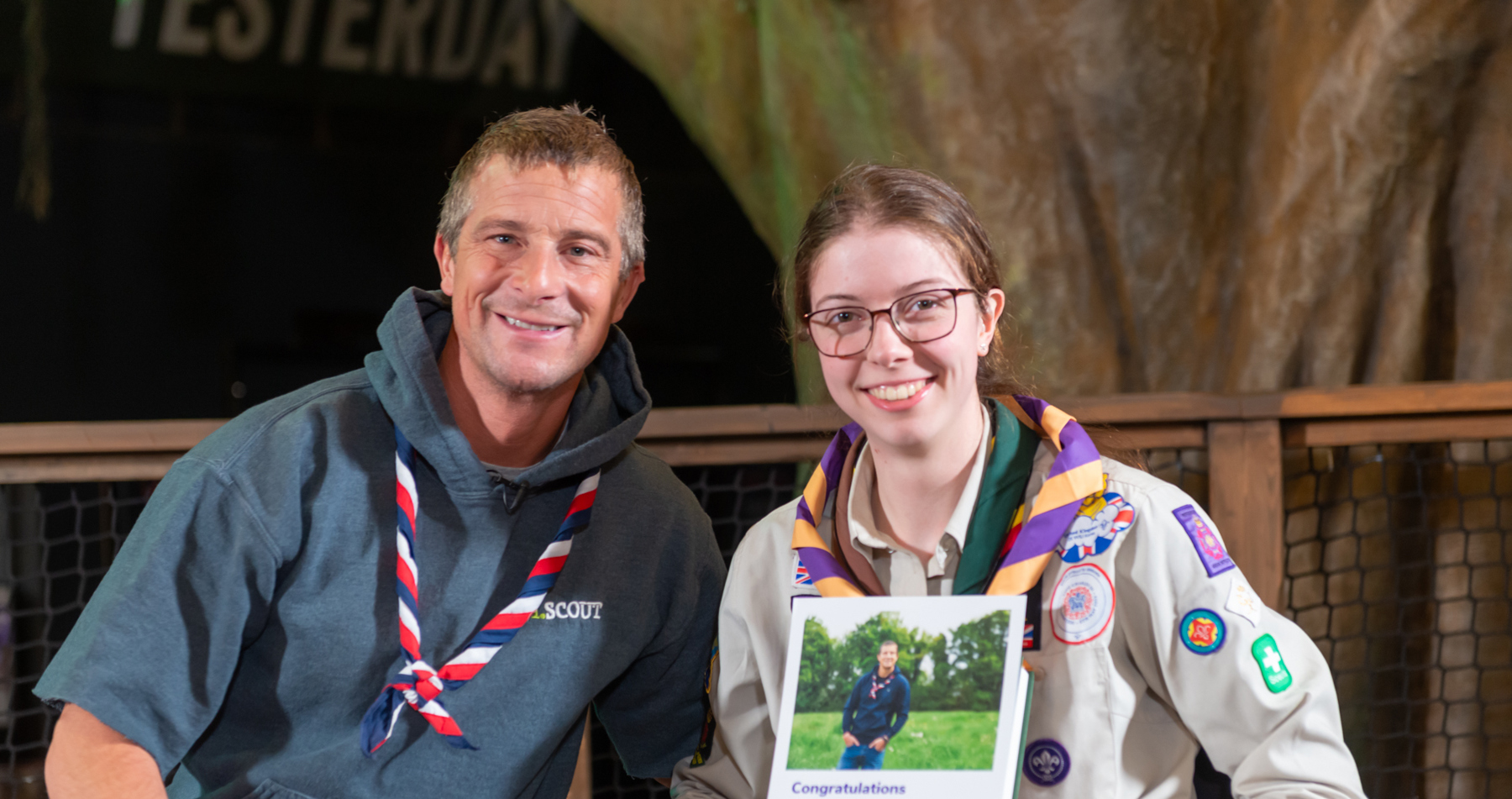 Chief Scout Bear Grylls is pictured with Unsung Hero award recipient Georgie. Bear is wearing a red, white and navy necker, and Georgie is stood to his right, wearing a purple and orange necker. Georgie's holding a certificate in her left hand. 