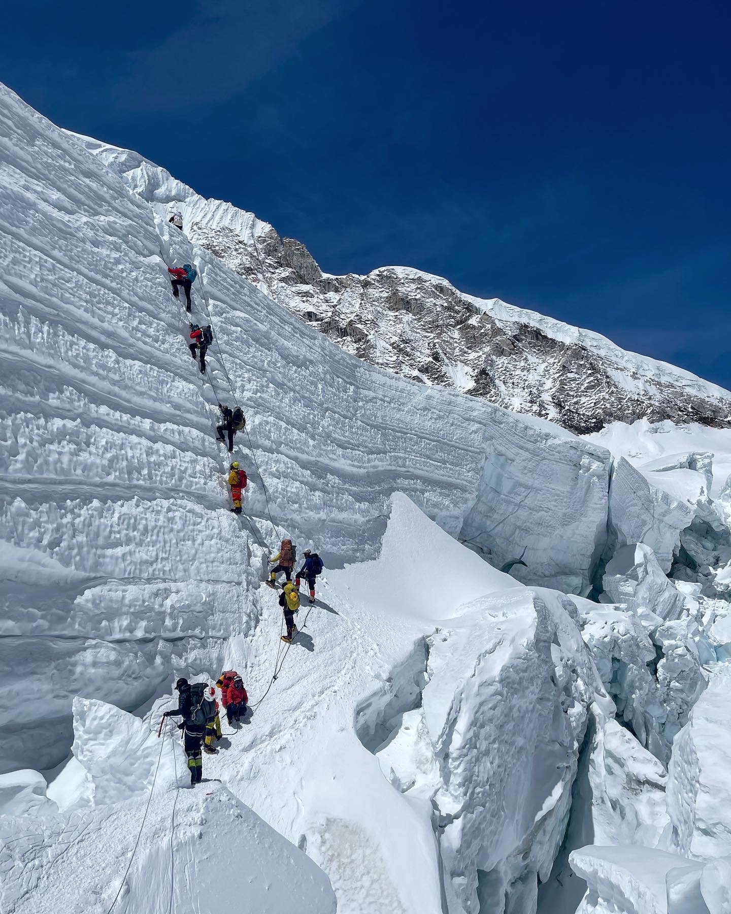 A group of mountaineers are climbing up a steep, snowy mountainside near Mount Everest. There are around 10 people waiting in line to climb up the rope and to the top of the mountain.