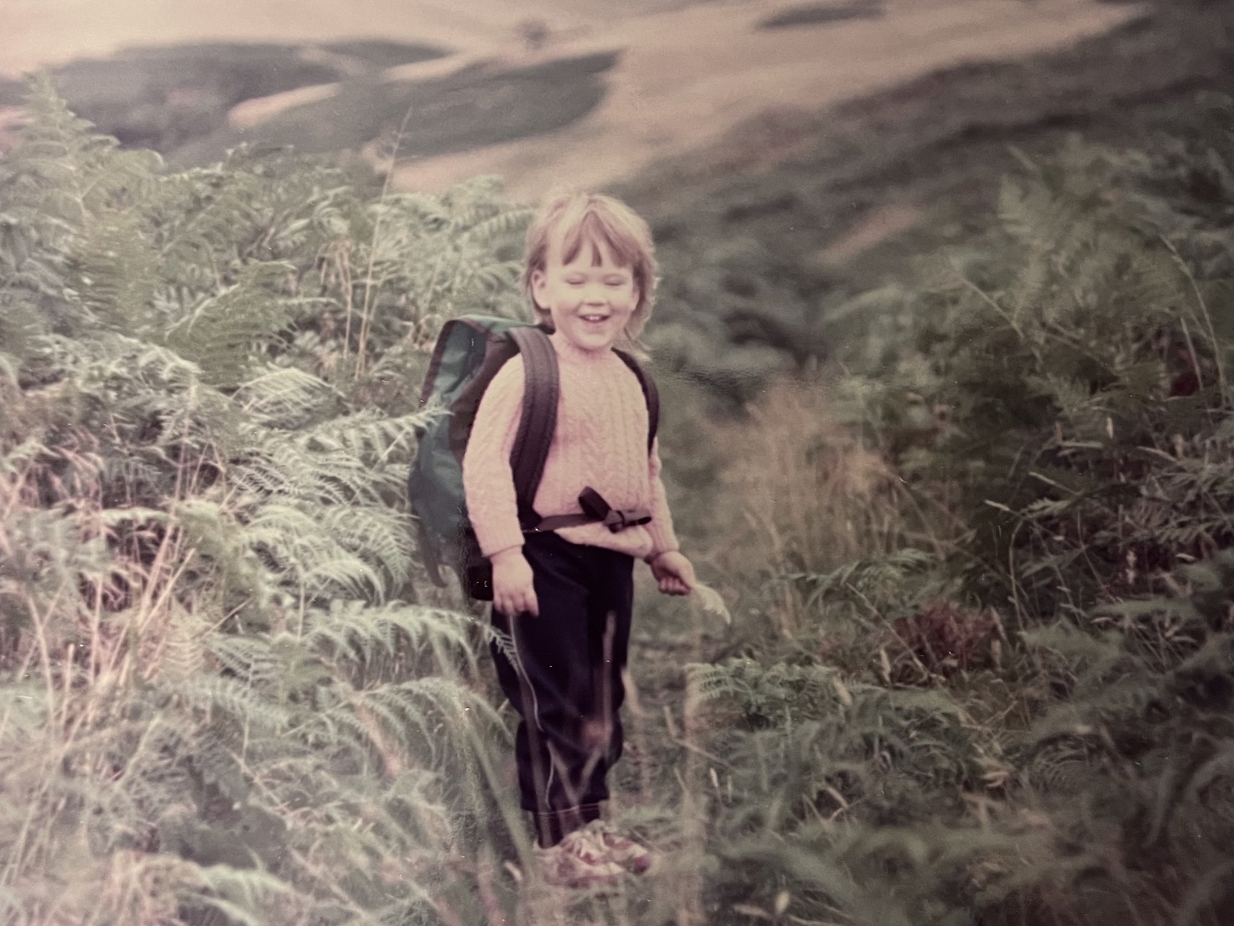 The image shows Kirsty Mack as a child, smiling with her eyes shut. She's stood in the wilderness with bushes around her, wearing a wool jumper, backpack, trousers and trainers.
