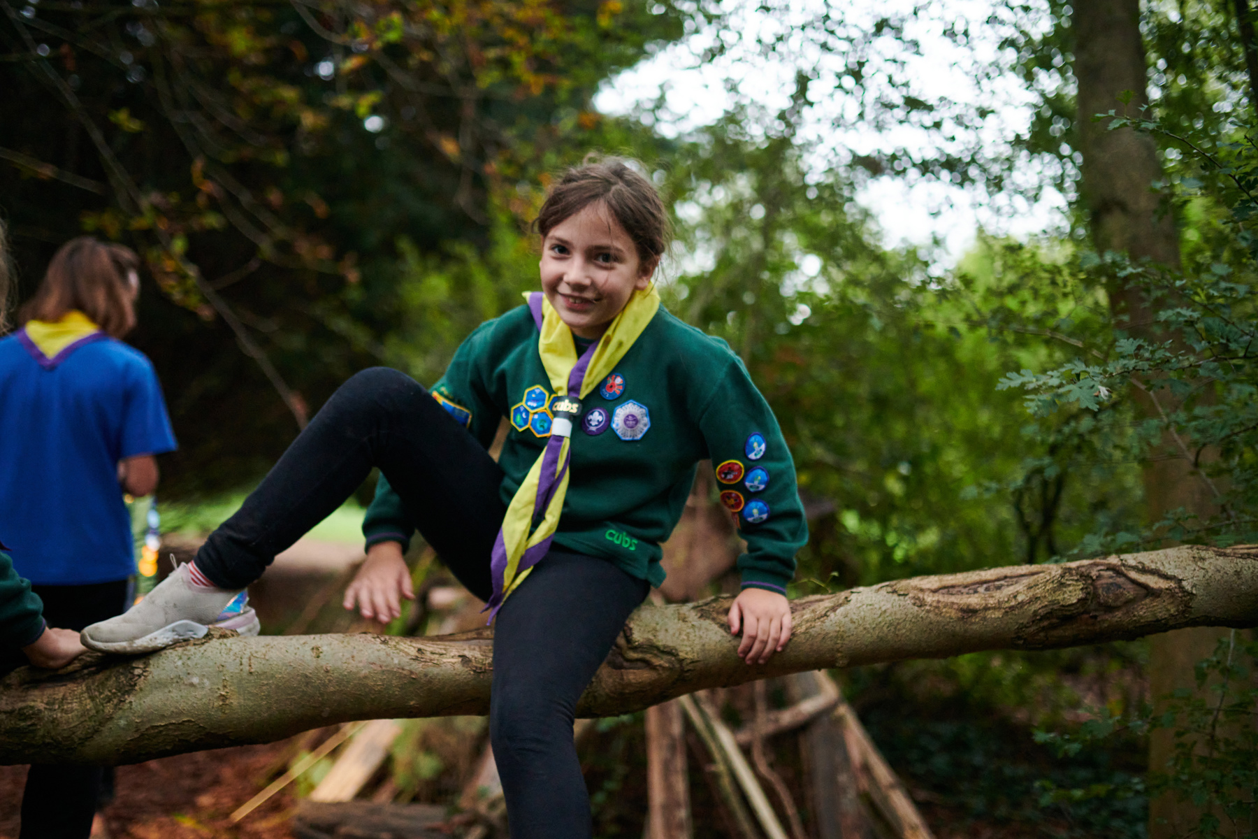 A smiling Cub in a green uniform and yellow and purple necker sits on a fallen tree and smiles.