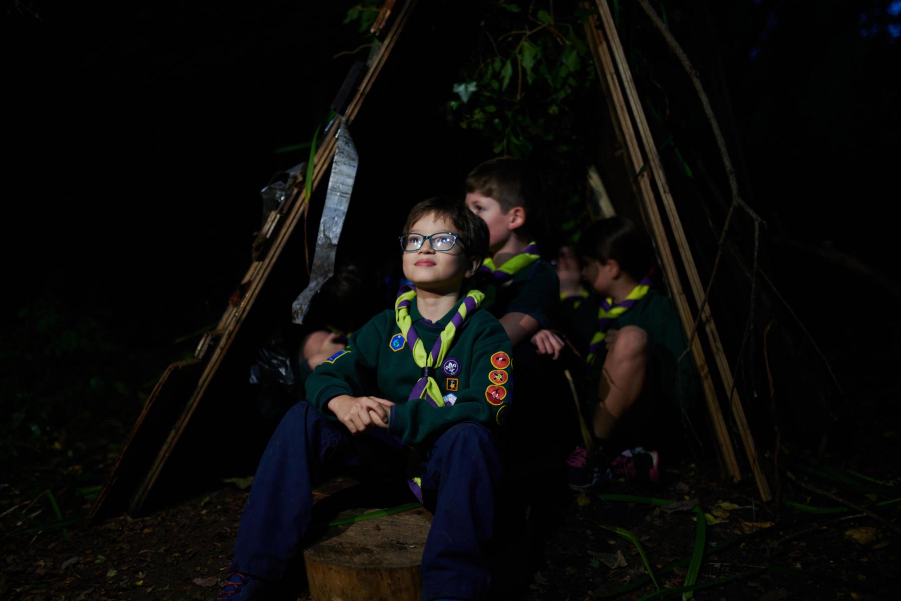 A boy in Cub uniform is seated outside a tent at night.
