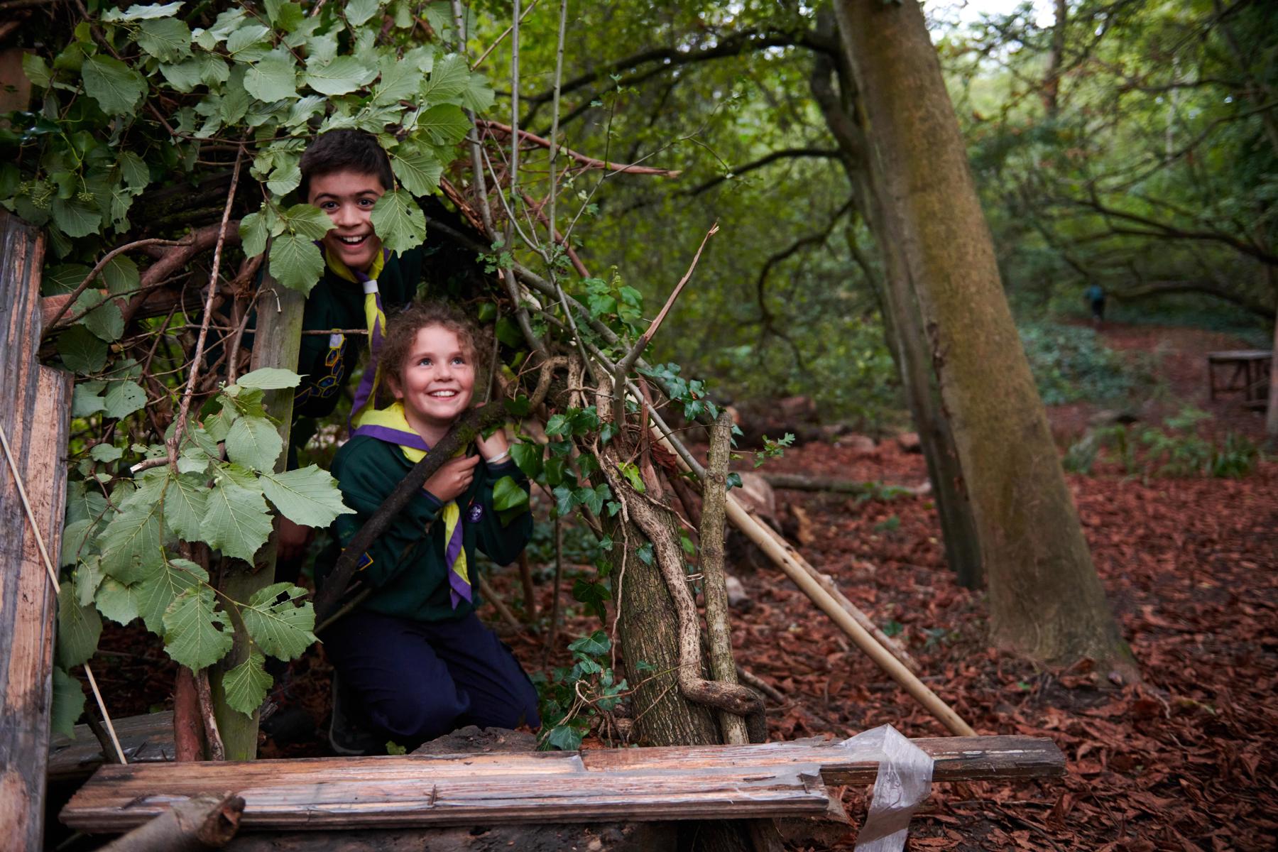 Two Cubs wearing green jumpers and yellow and purple scarves smile while sat inside a den.