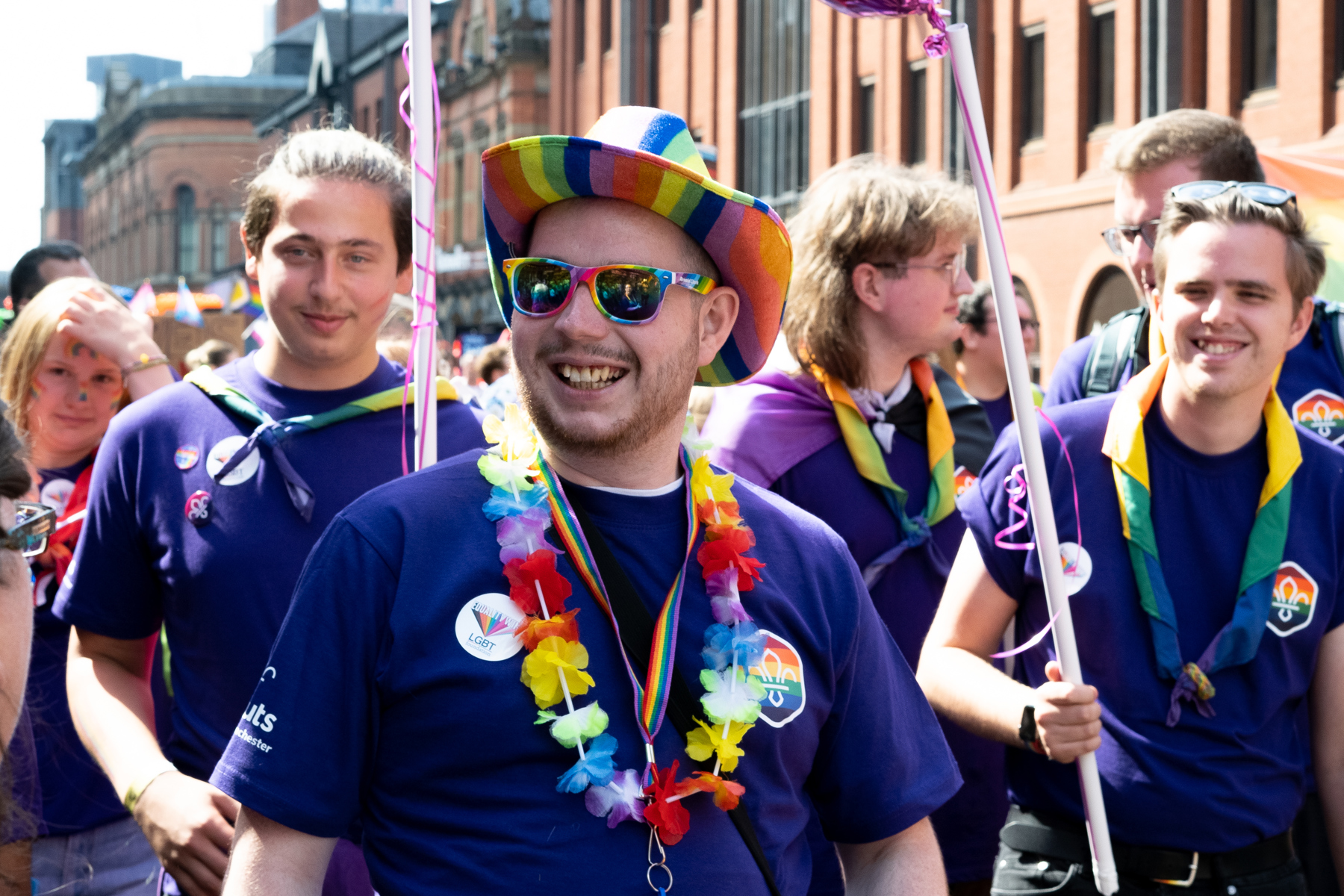 A Scout in a Pride parade wearing a rainbow necker, rainbow hat, rainbow sunglasses and Scout Pride t-shirt. They are smiling.