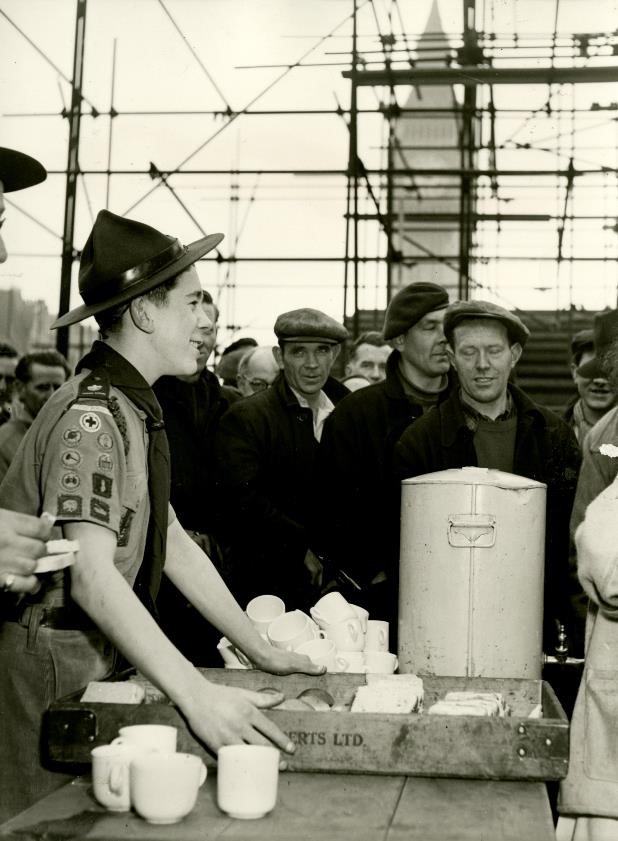 Black and white photo of Scout holding a tray of tea cups with men in flat caps standing around waiting for a cup of tea during the build up of the 1953 Coronation