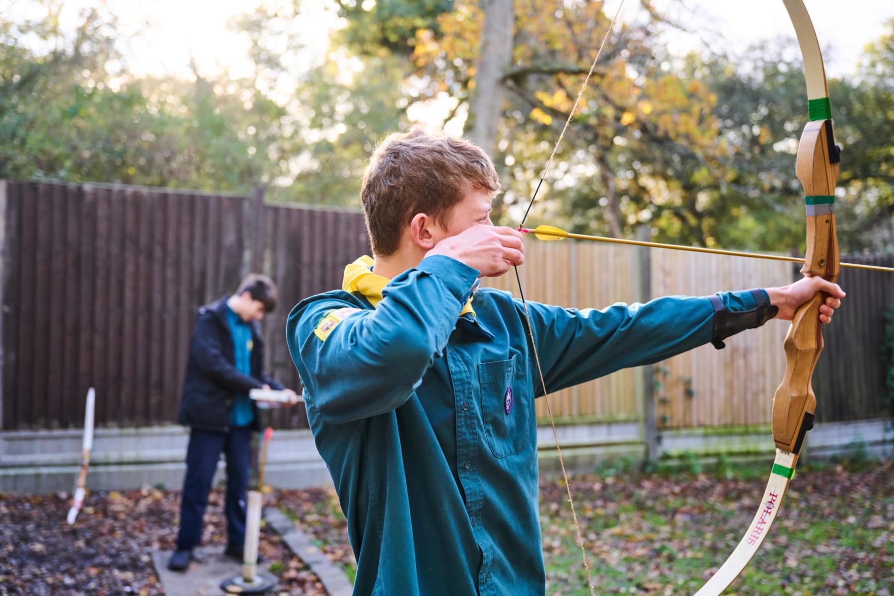 A Scout in uniform holds up a bow and arrow taking part in archery outside.