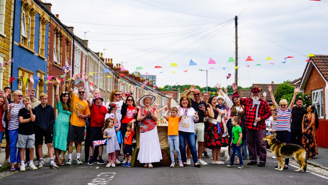 Crowd of people in brightly coloured clothes standing in the middle of a bunting-lined street waving at the camera