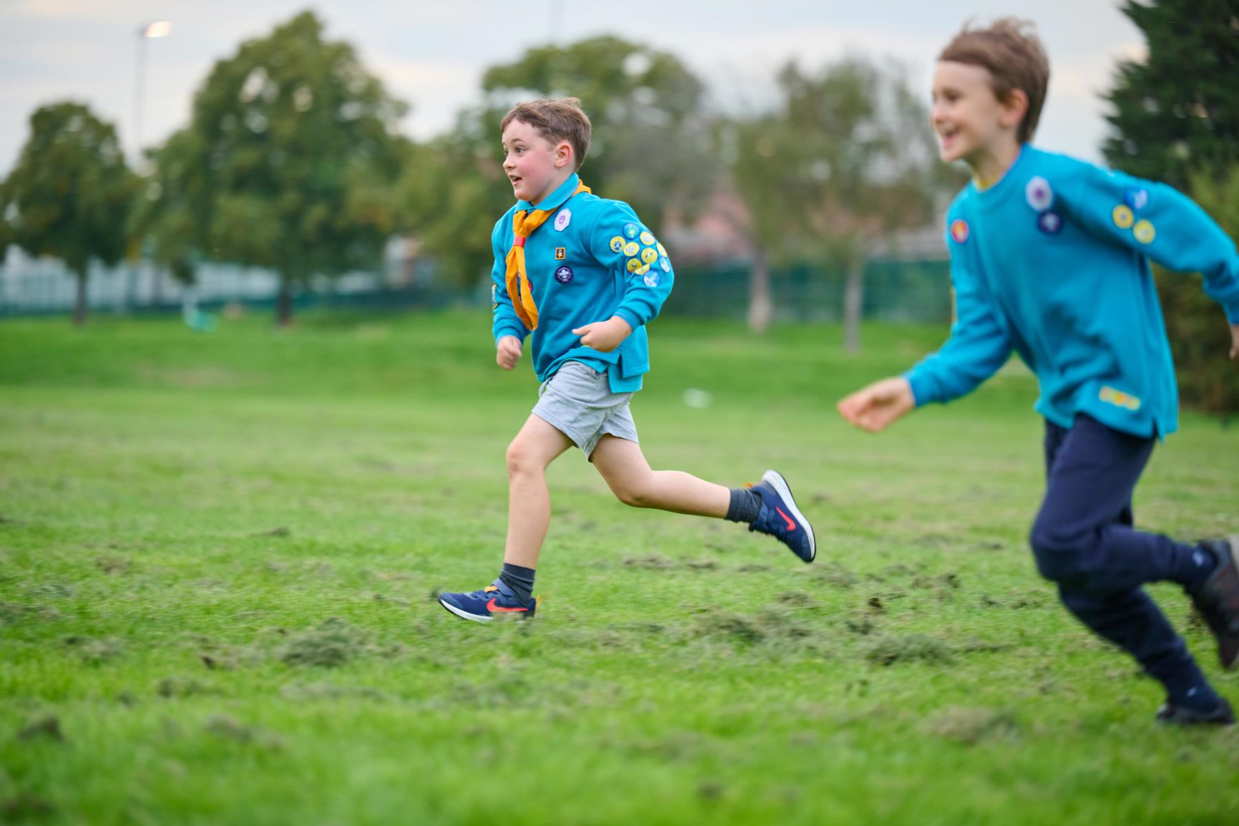 Two male Beavers running across a field. They are wearing blue jumpers, one is wearing a yellow neck. They are both smiling.