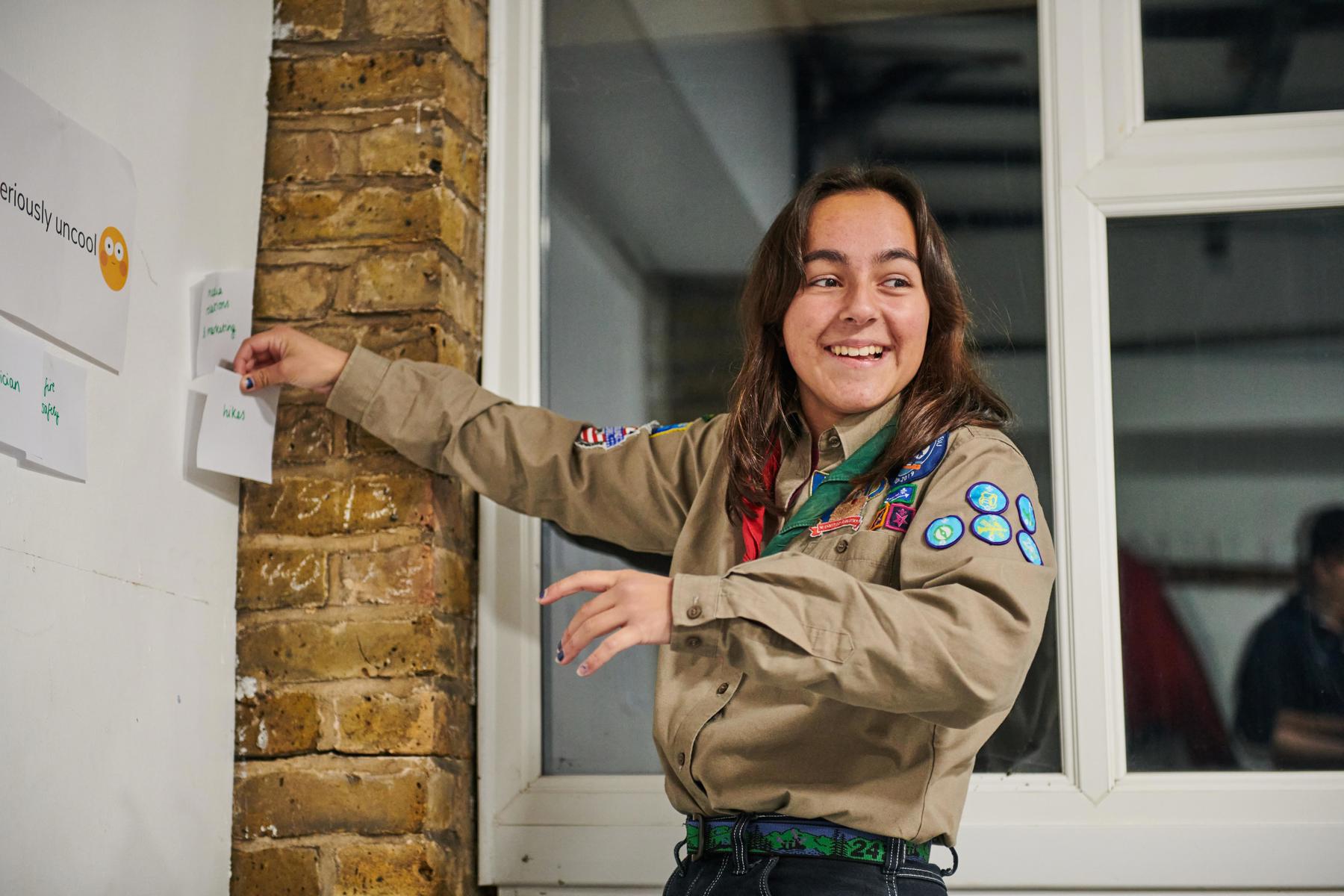 An Explorer wearing their uniform and necker is smiling to someone off camera as they stick a note to a whiteboard.