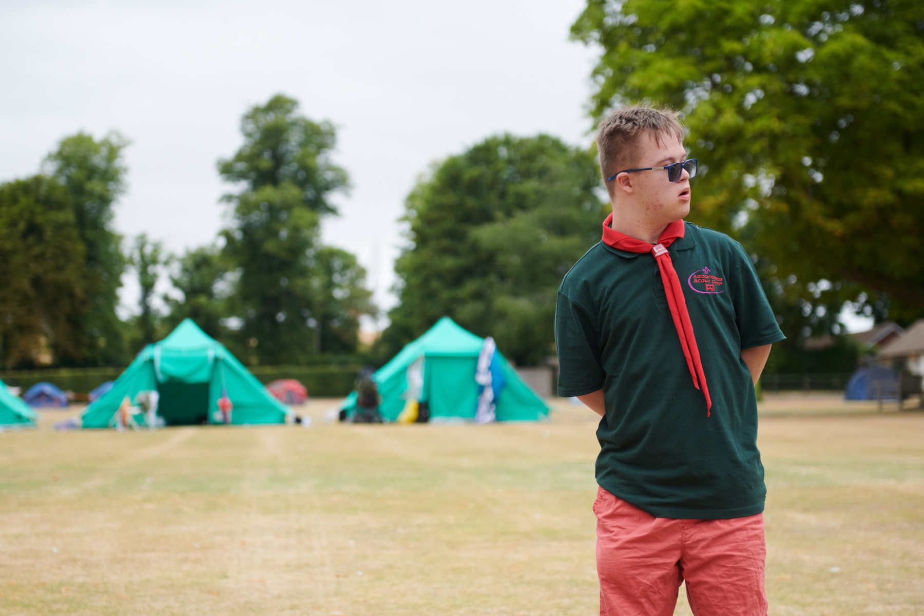 George is stood in a field on camp. There are tents and trees behind him. He's in a green Scouts top, red necker and sunglasses. He's looking over to the right.