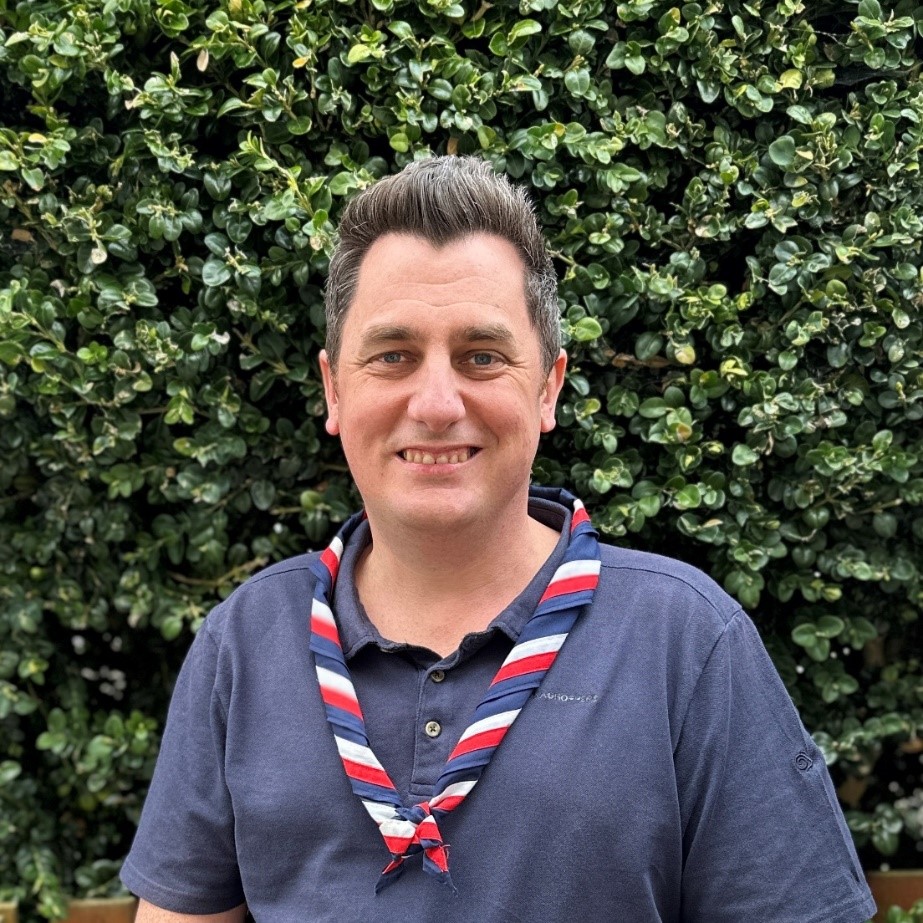 Barry Donald-Hewitt smiling at the camera while wearing a navy Scouts polo shirt and stripy scarf