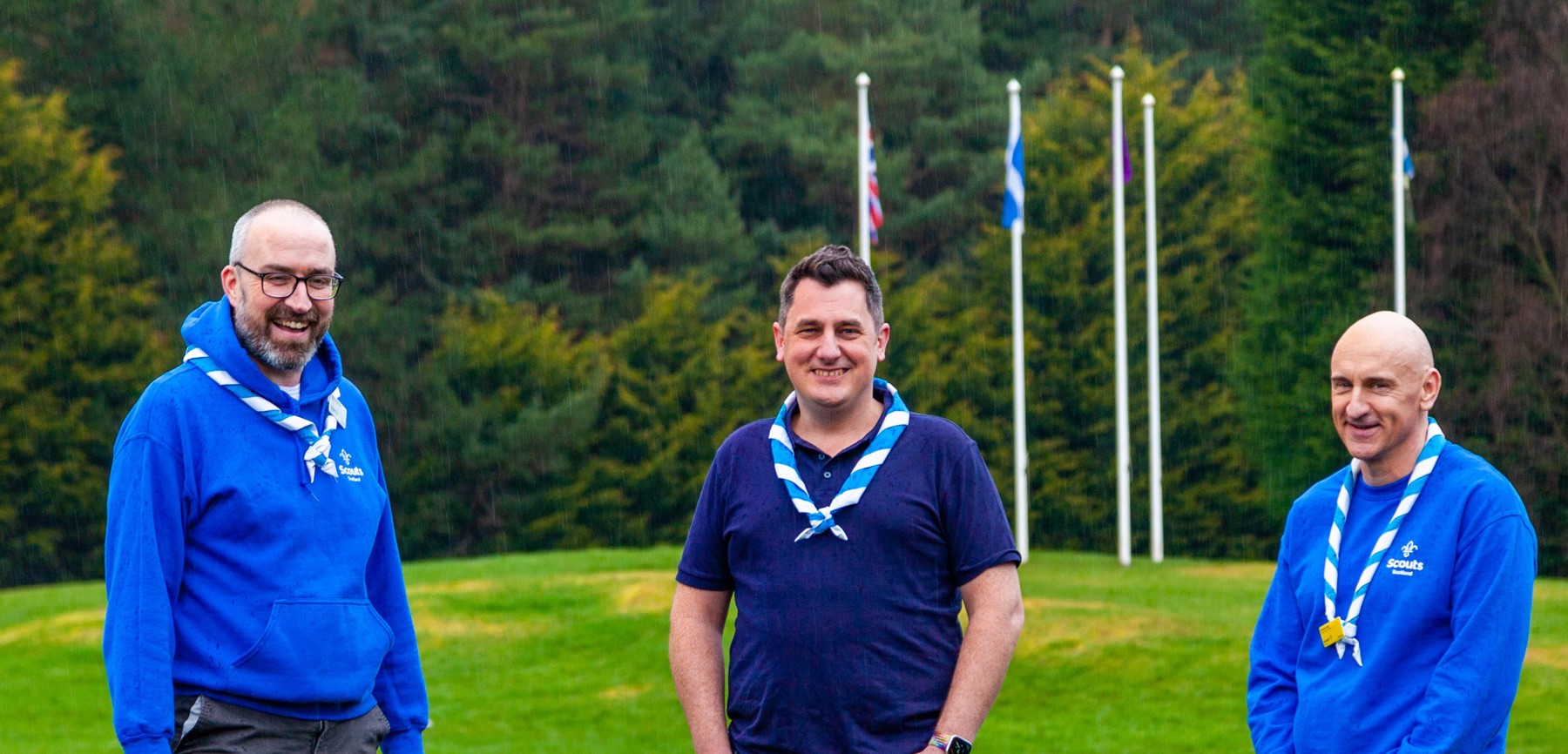 Three men standing in front of the UK and Scotland flags wearing blue and blue and white neckers