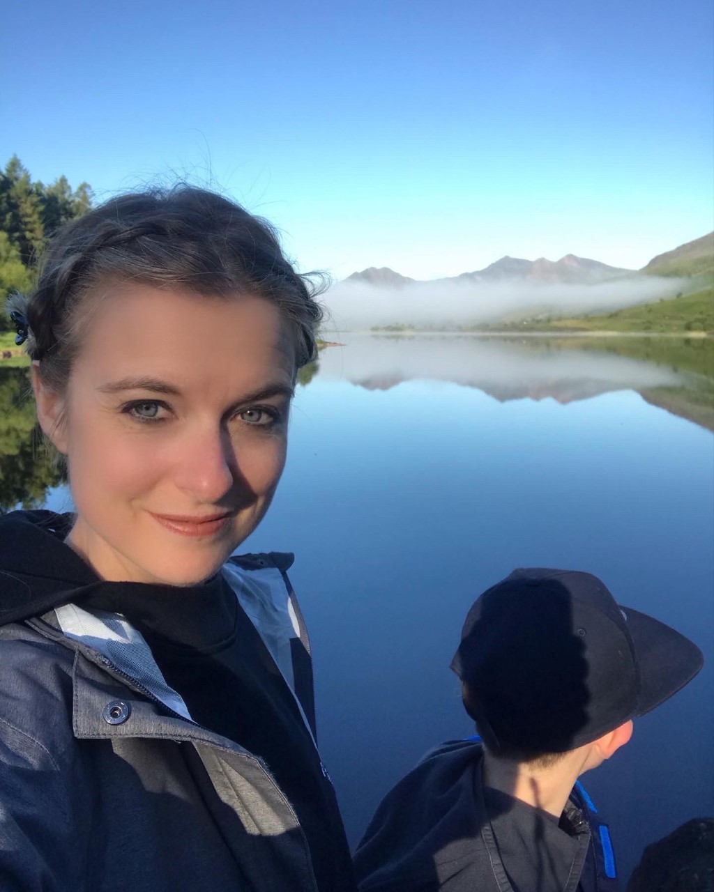 Lucy Cox stands by a lake with mountains in the background.