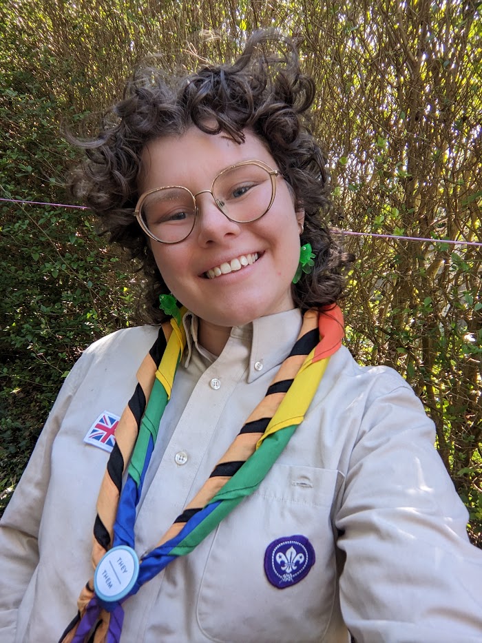 In their volunteer uniform, Anna is smiling. Anna wears glasses and a 'They/Them' badge.