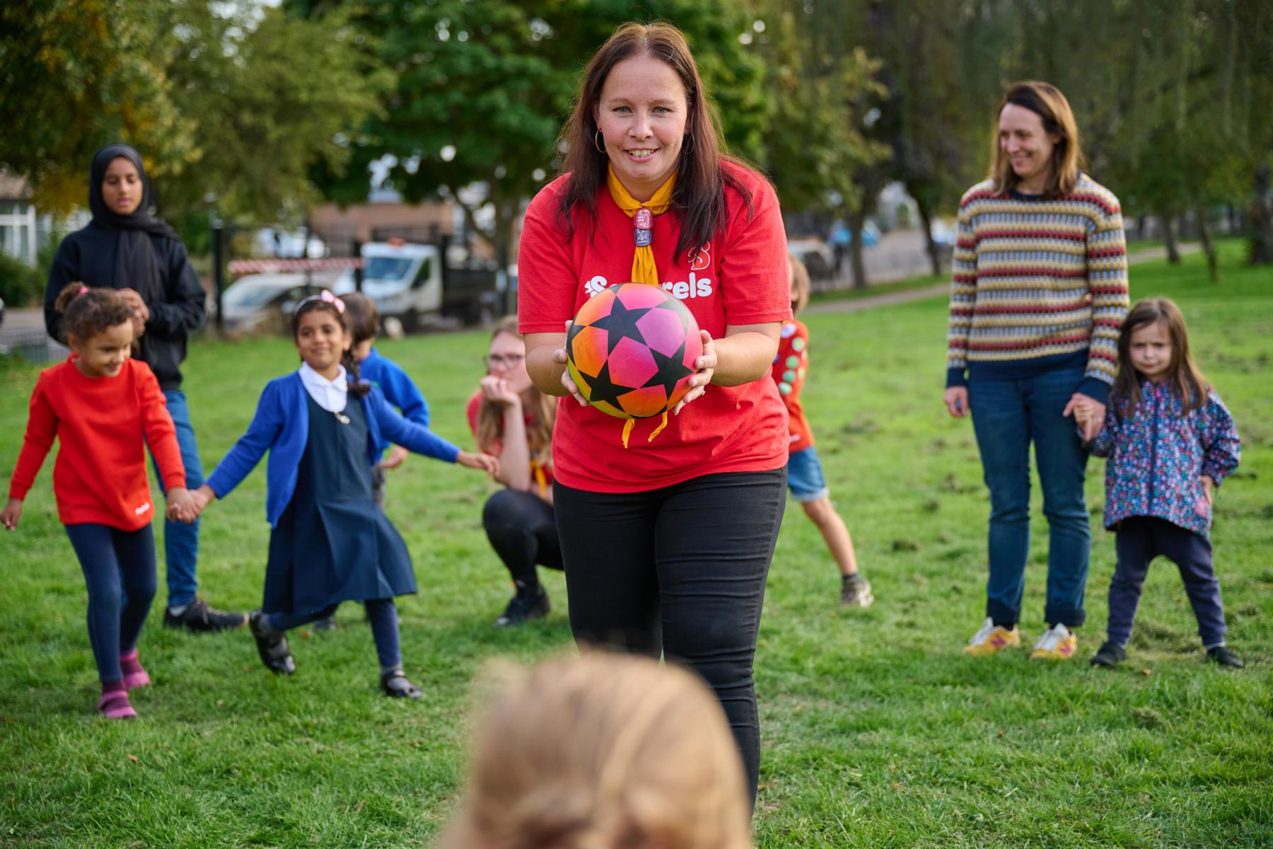 A volunteer wearing a red Squirrels t-shirt holds a ball in their hands while standing in a park with a group of young people.