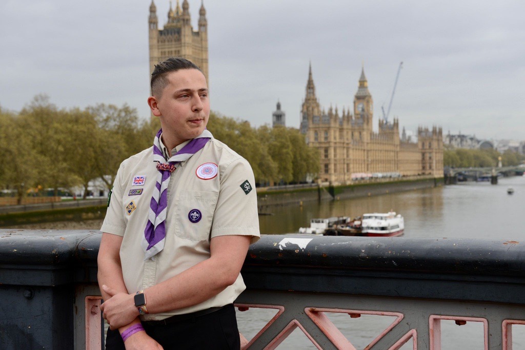 A Scout wearing uniform stood on a bridge in London by the Houses of Parliament