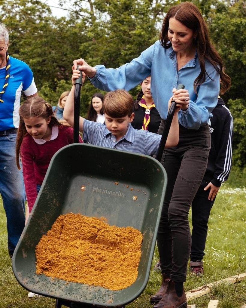 Princess Charlotte, Prince Louis and the Princess of Wales are all holding and pushing a wheelbarrow full of sand.
