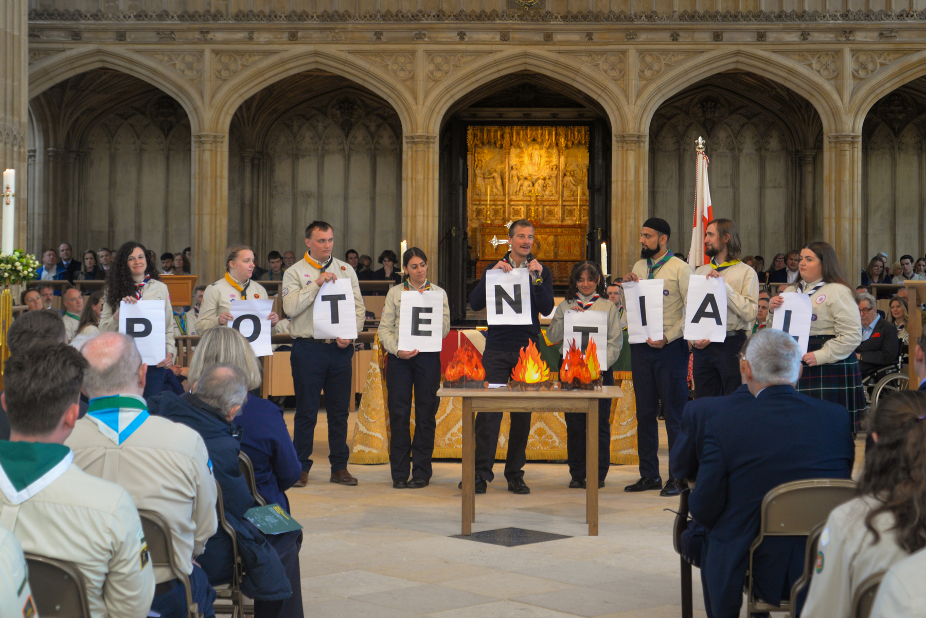 Bear Grylls is standing in front of a row of Scouts in uniform inside St George's Chapel. Each Scout and Bear is holding a piece of paper with a letter on, spelling out the word 'POTENTIAL.'