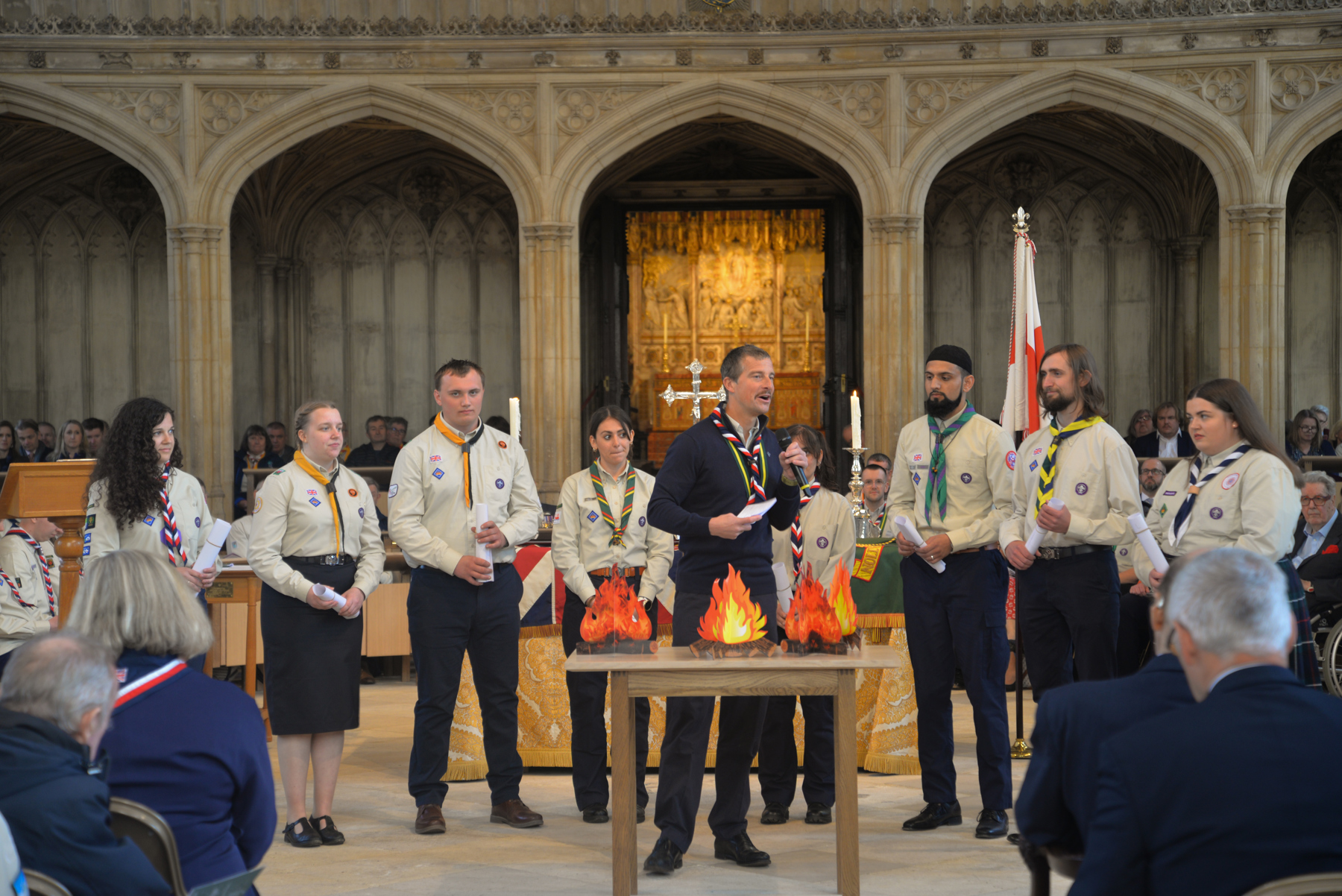 Chief Scout Bear Grylls is standing in a chapel holding a microphone as he gives a speech. Stood behind him are a row of Scouts in uniforms and neckers.