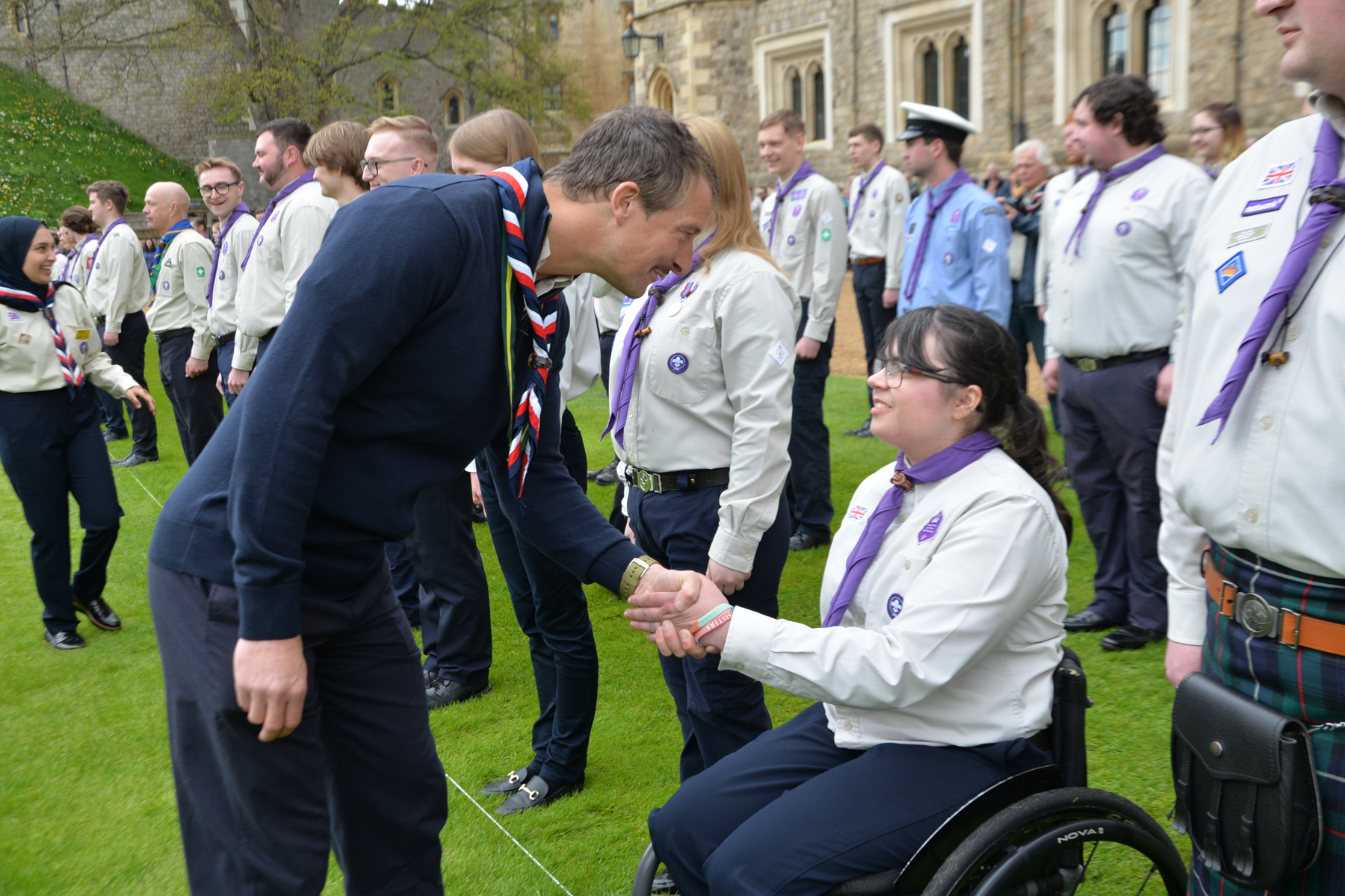 Bear Grylls is wearing a necker and standing on grass. He's bending over to shake the hand of a Scout in uniform who's in a wheelchair. Behind them are rows of other Scouts also in uniforms and neckers.