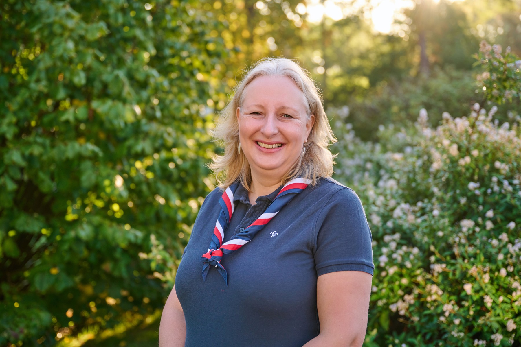 Kate Marks, Scouts Trustee, stands in front of bushes with sunlight behind her. She's smiling at the camera and she's wearing a navy Scouts polo shirt with a red, blue and white necker.
