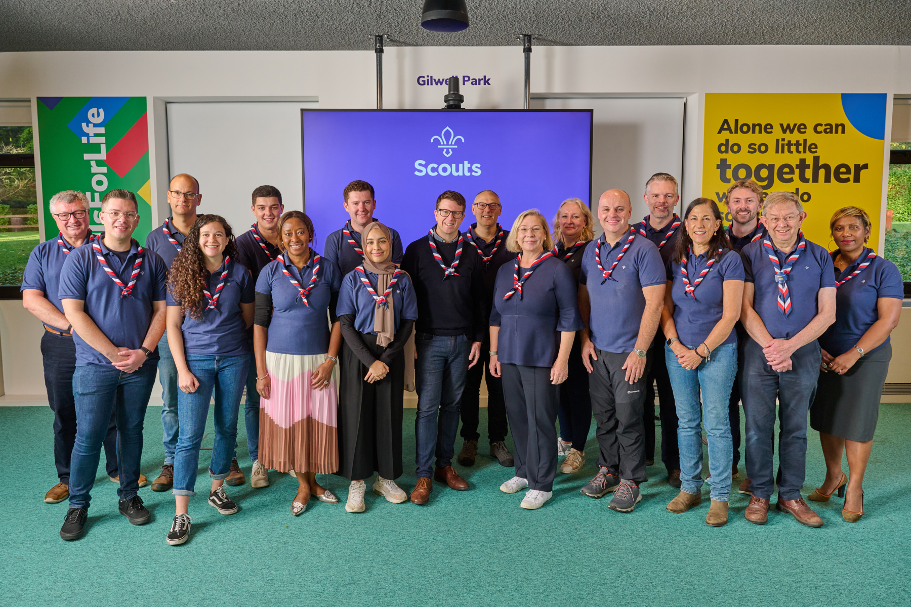 The Scouts Board of Trustees stand together in a line, all wearing navy polo shirts and red, white and blue neckers. They're smiling at the camera and standing in front of a TV screen showing the Scouts logo in white on a purple background.
