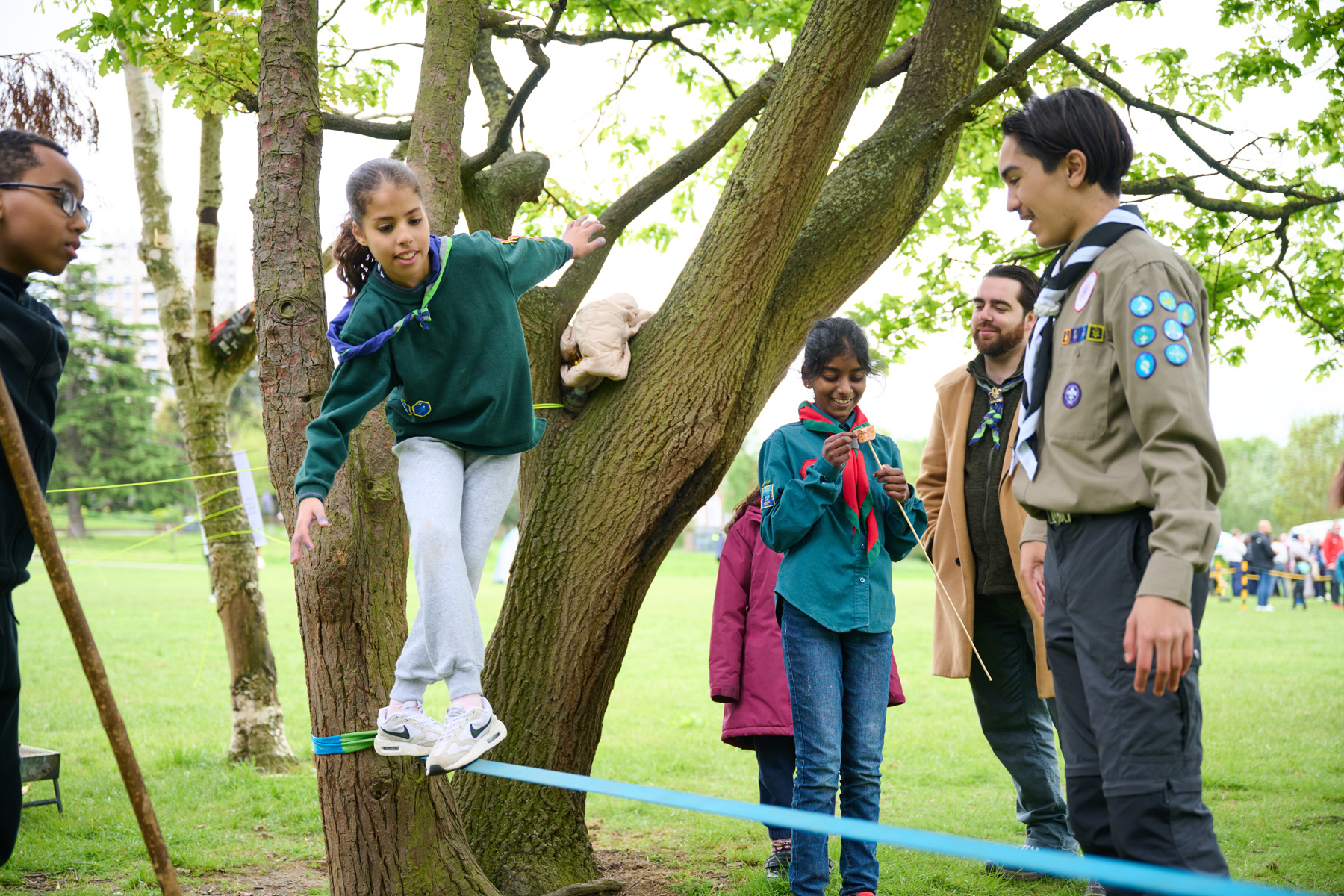 A Cub is standing on a blue rope tied to a tree and is trying to balance on it. A Scout, two volunteers and another Cub are stood next to the rope as she stands on it.