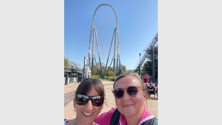 Two women are stood close to the camera and you can just see their heads. They're both wearing sunglasses and are smiling at the camera. They're stood in front of a very tall rollercoaster ride and there's no clouds in the bright blue sky.