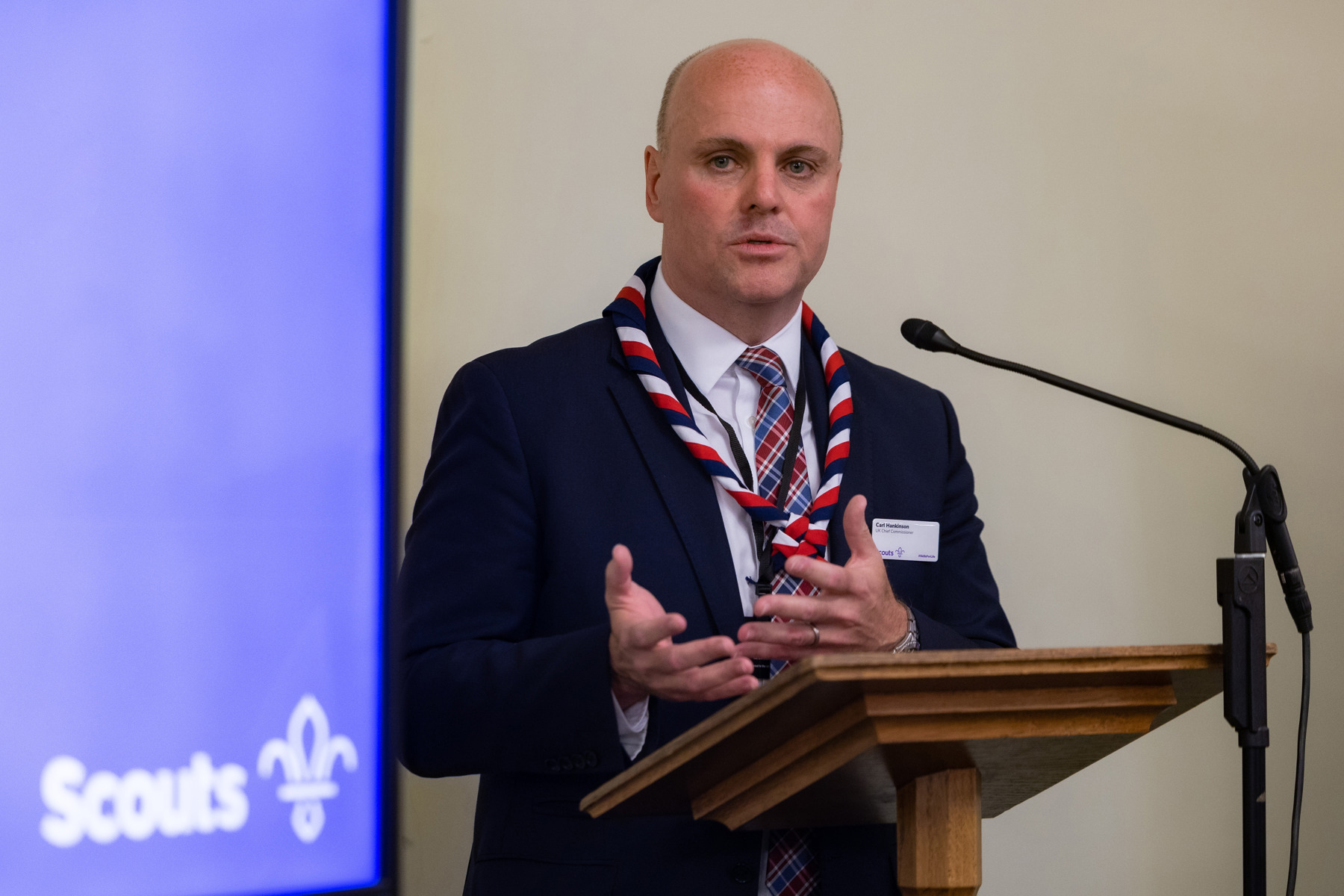 UK Chief Commissioner Carl Hankinson is wearing a suit and necker as he stands in front of a microphone to present at Parliament. In the background to the left is a purple screen with a white Scouts logo on.